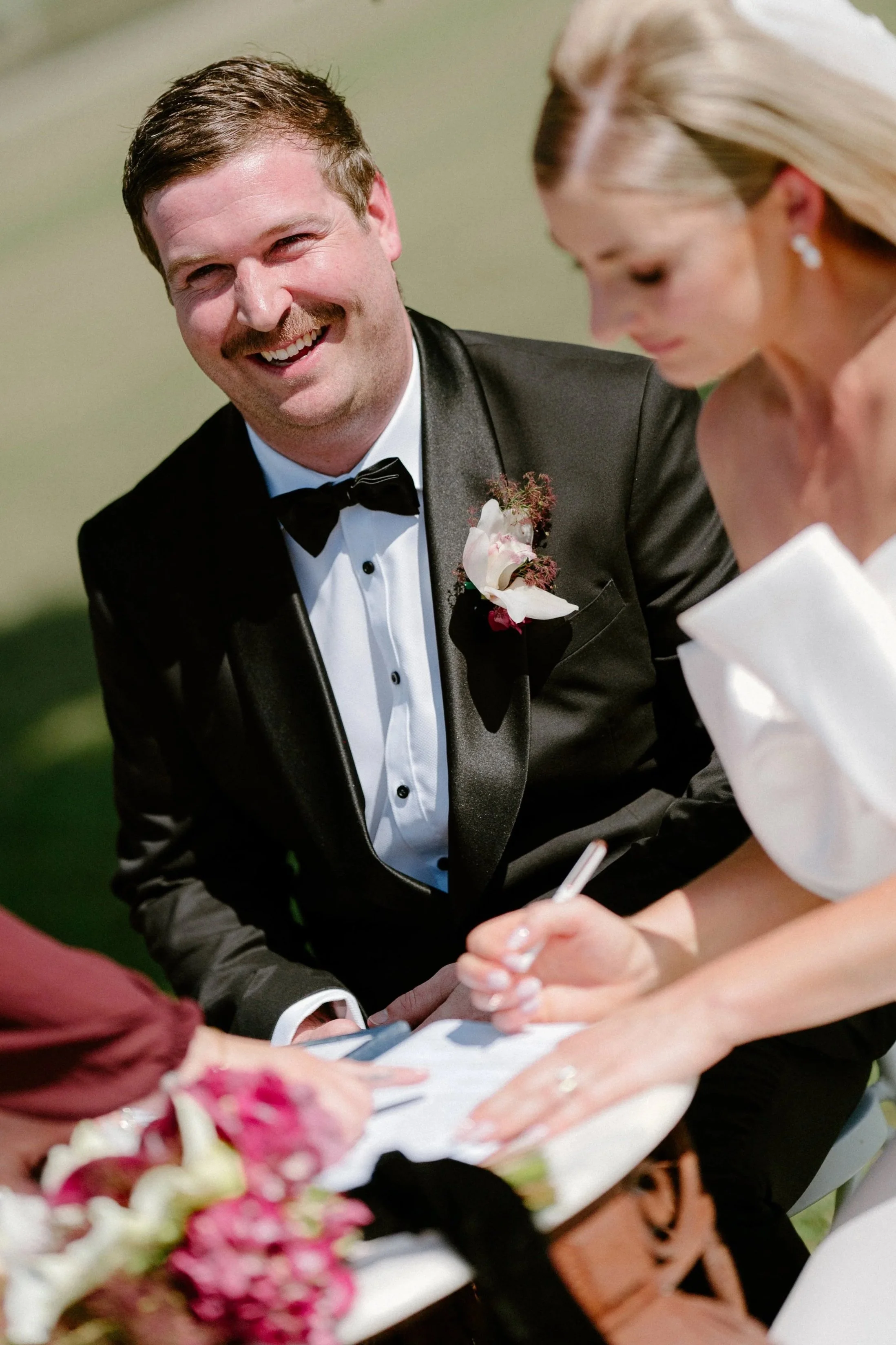 A smiling groom in a black tuxedo with a boutonniere signs a marriage certificate with a pen, seated outdoors at a wedding ceremony.