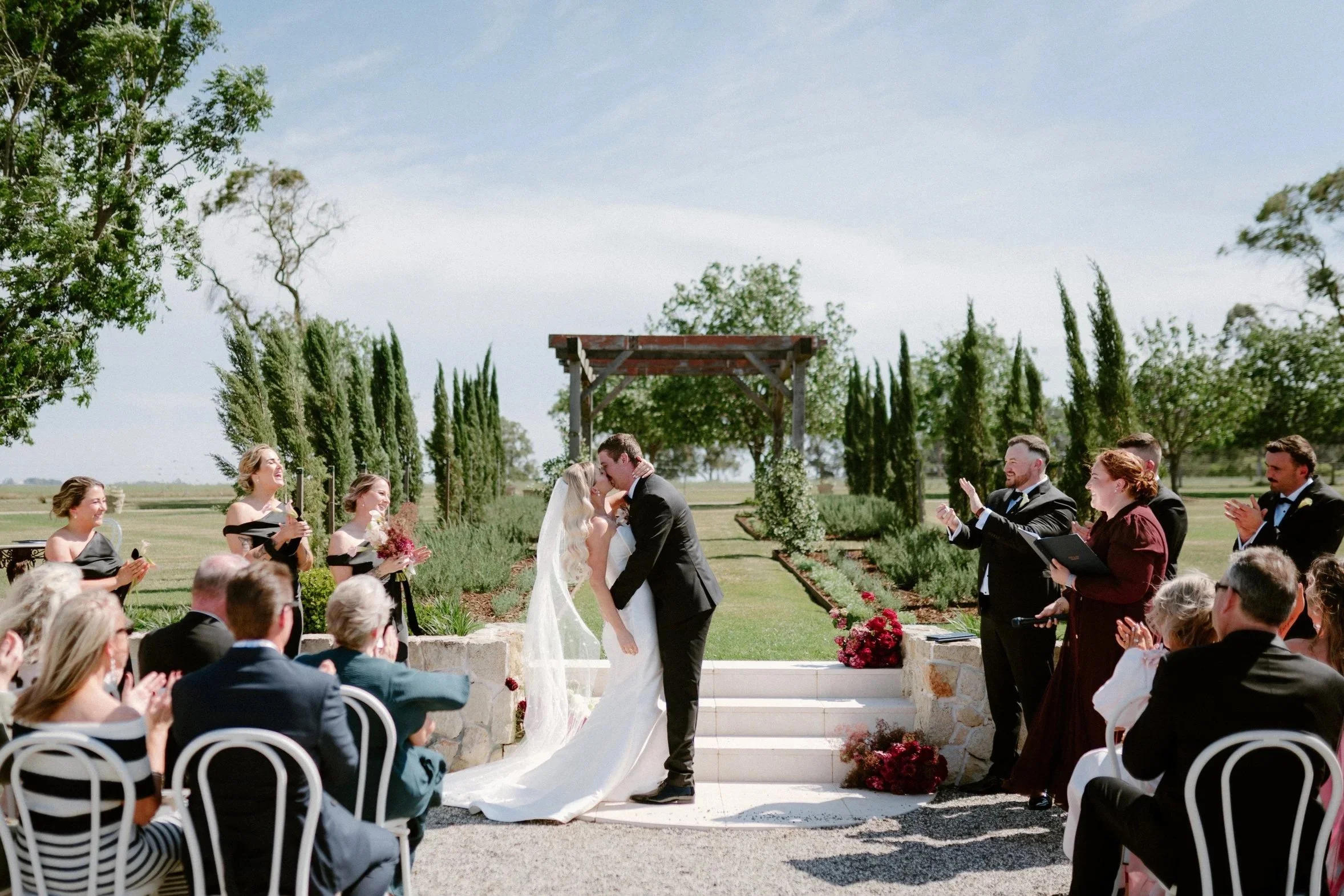 A wedding ceremony taking place outdoors with a bride and groom kissing at the altar, surrounded by seated guests, on a sunny day with blue skies and green trees.