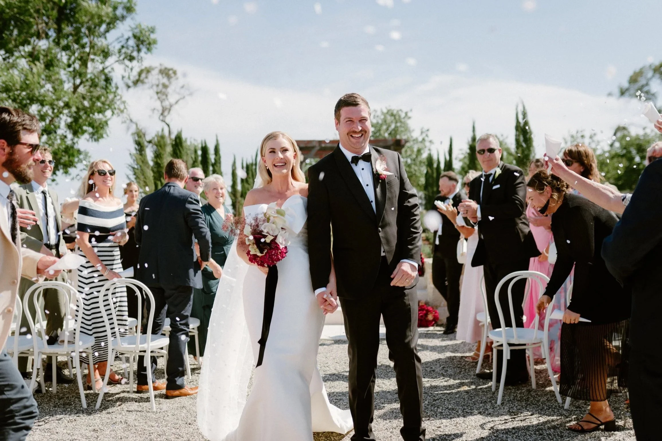 A newly married couple walks hand in hand down the aisle, smiling, as guests celebrate by tossing confetti. The bride wears a white off-the-shoulder wedding gown and holds a bouquet, while the groom is in a black tuxedo. Guests are dressed in formal 