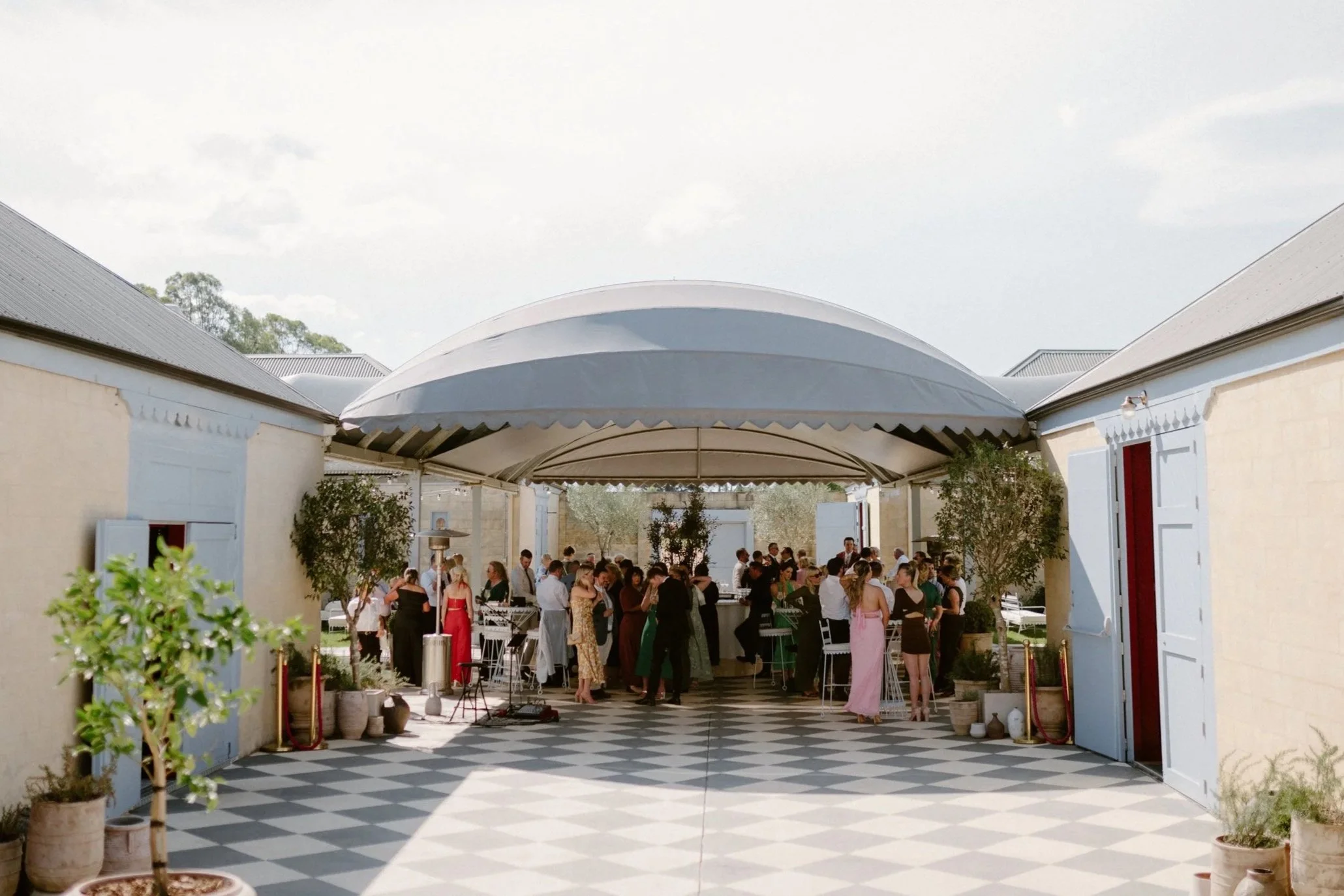 The Sienna, Hunter Valley Wedding Venue, Wedding Photos. People gathered at an outdoor event under a large white canopy, with a checkered black-and-white floor and potted plants along the sides.