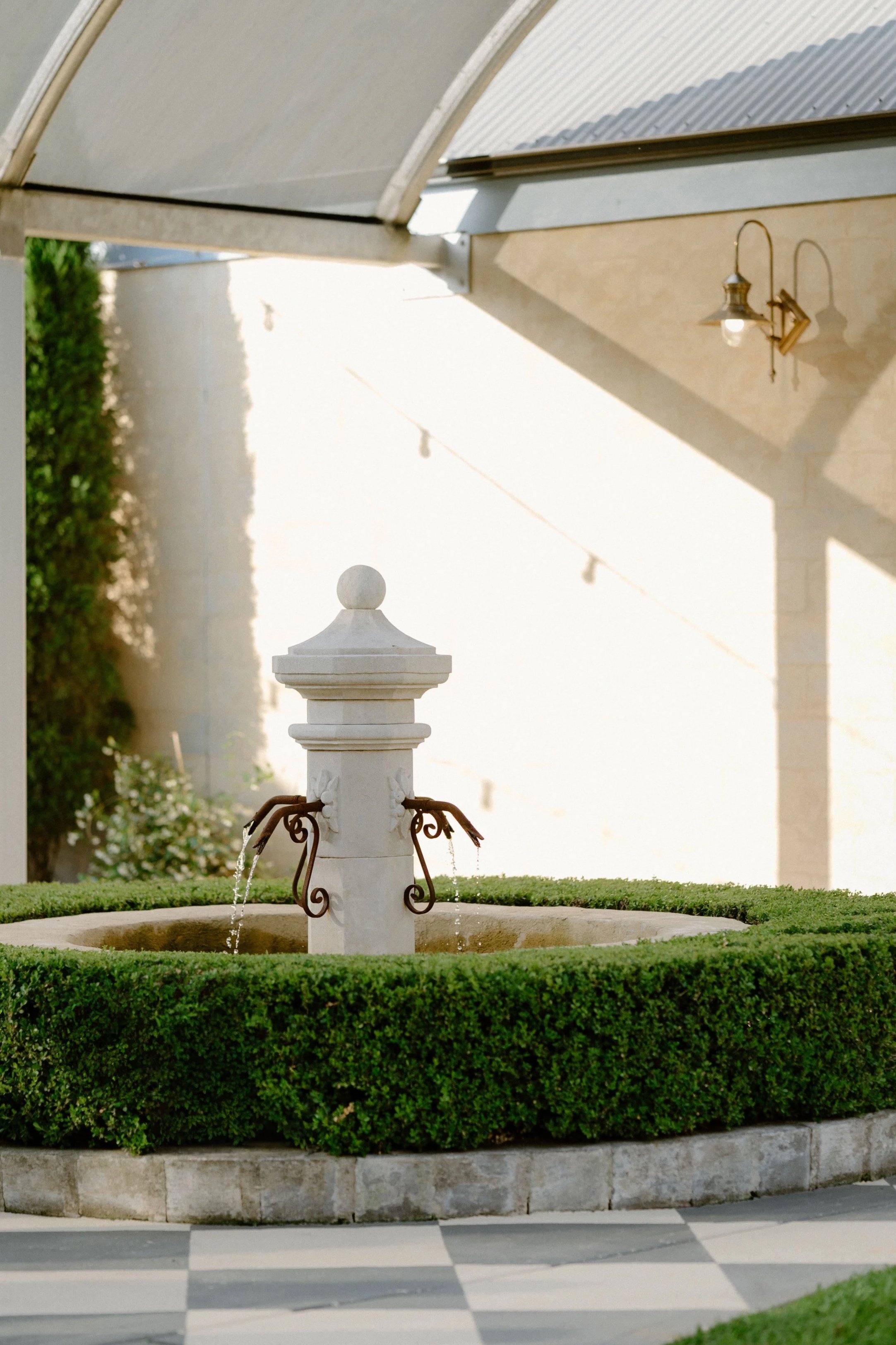 A small decorative fountain in an outdoor patio area, surrounded by a neatly trimmed hedge, with checkered black and white flooring, a covered patio roof, and a wall-mounted outdoor light.