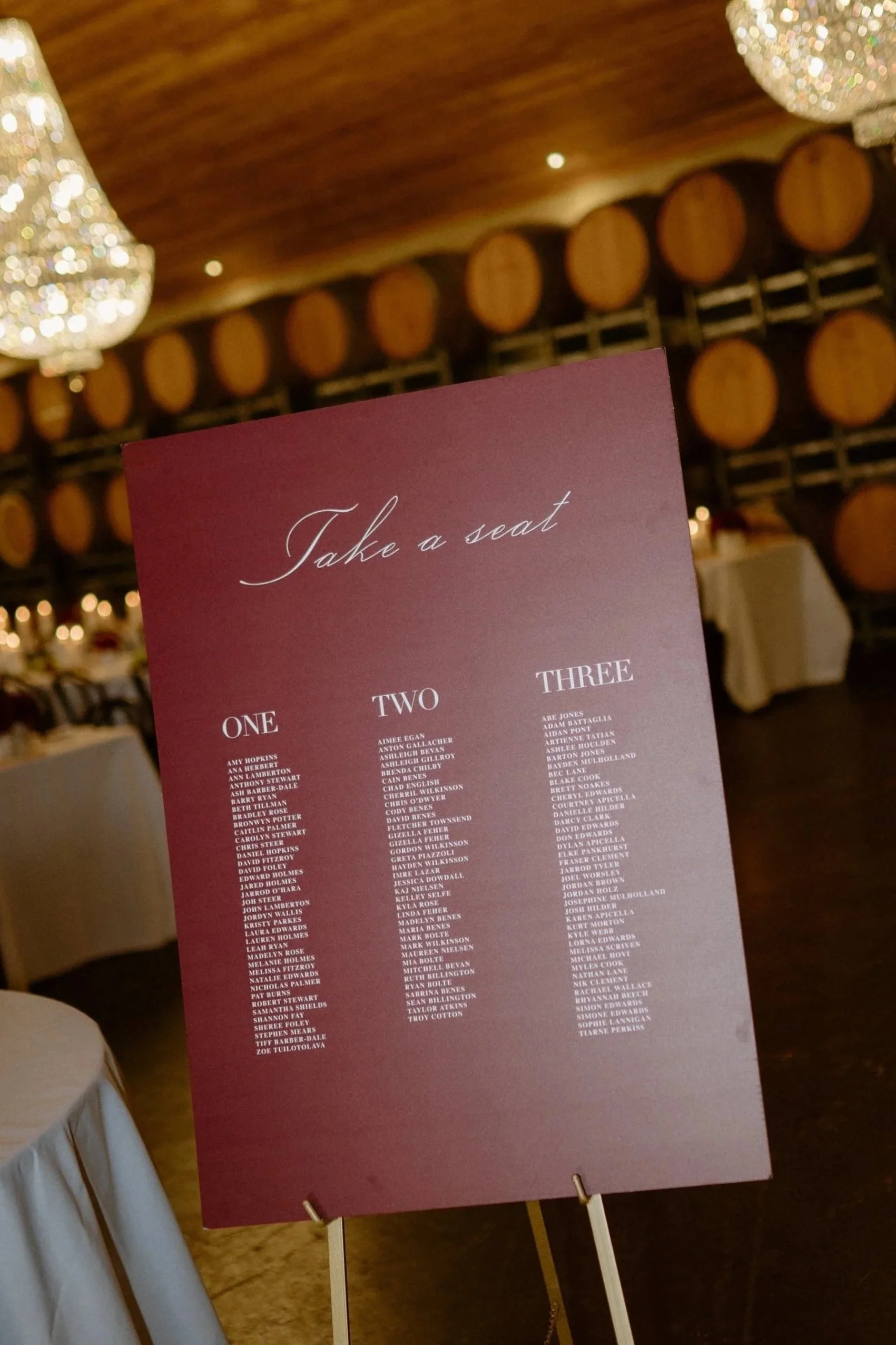 A wedding seating chart on a purple board displaying guest names in three columns, with the words 'Take a seat' at the top, set up on a stand inside a decorated venue with chandeliers and barrels in the background.