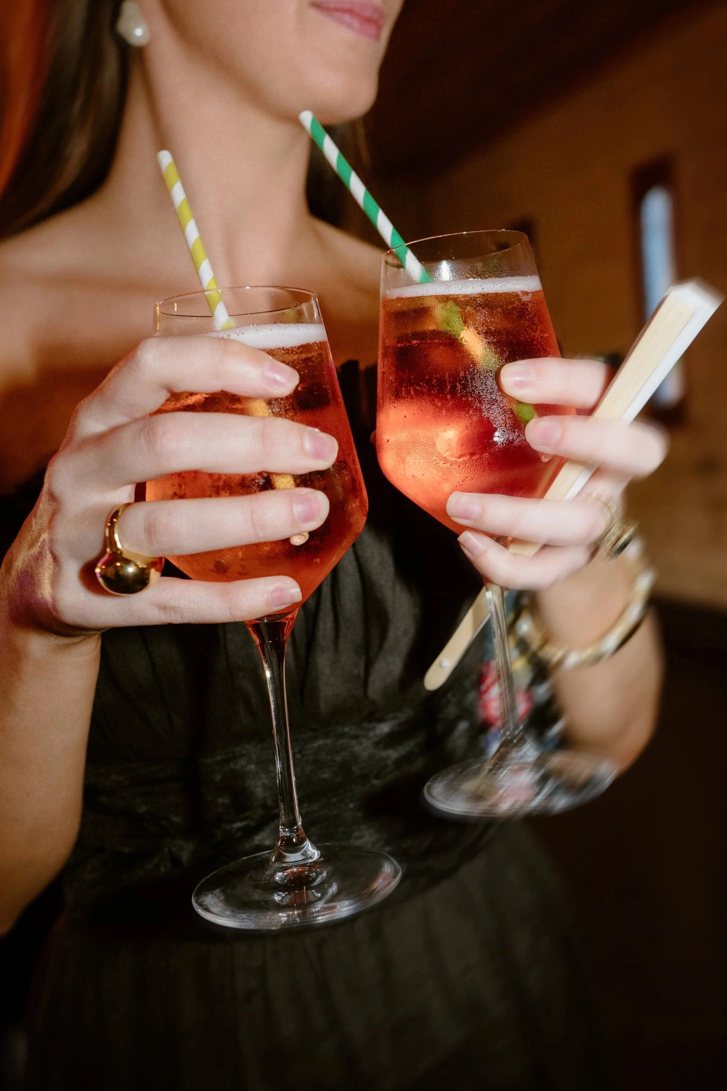 A woman holding two glasses of pink cocktails with striped straws, one with a lime wedge, in a bar or restaurant setting.