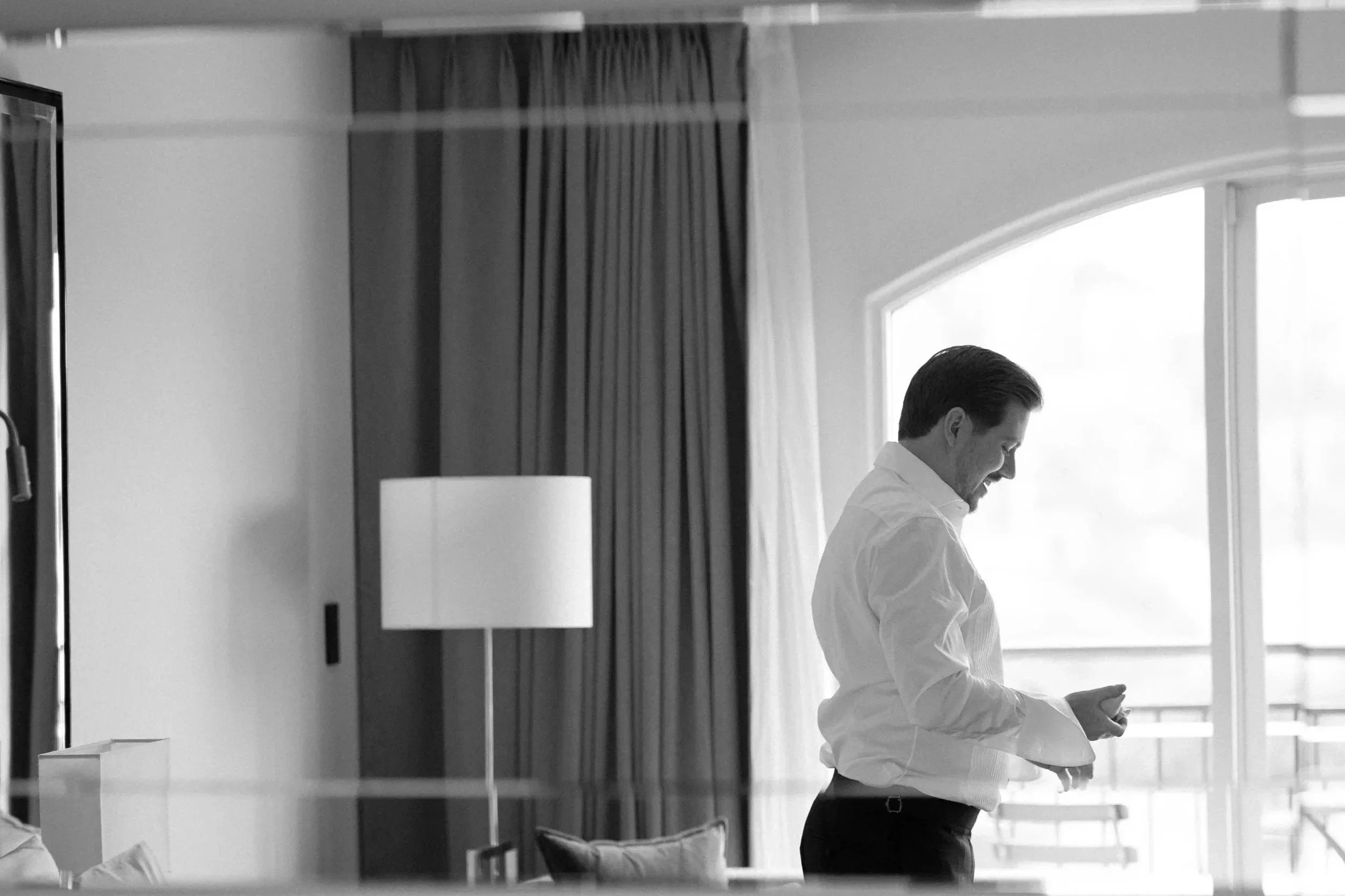 A man in a white shirt standing indoors near a large window with curtains, looking at his phone and smiling.
