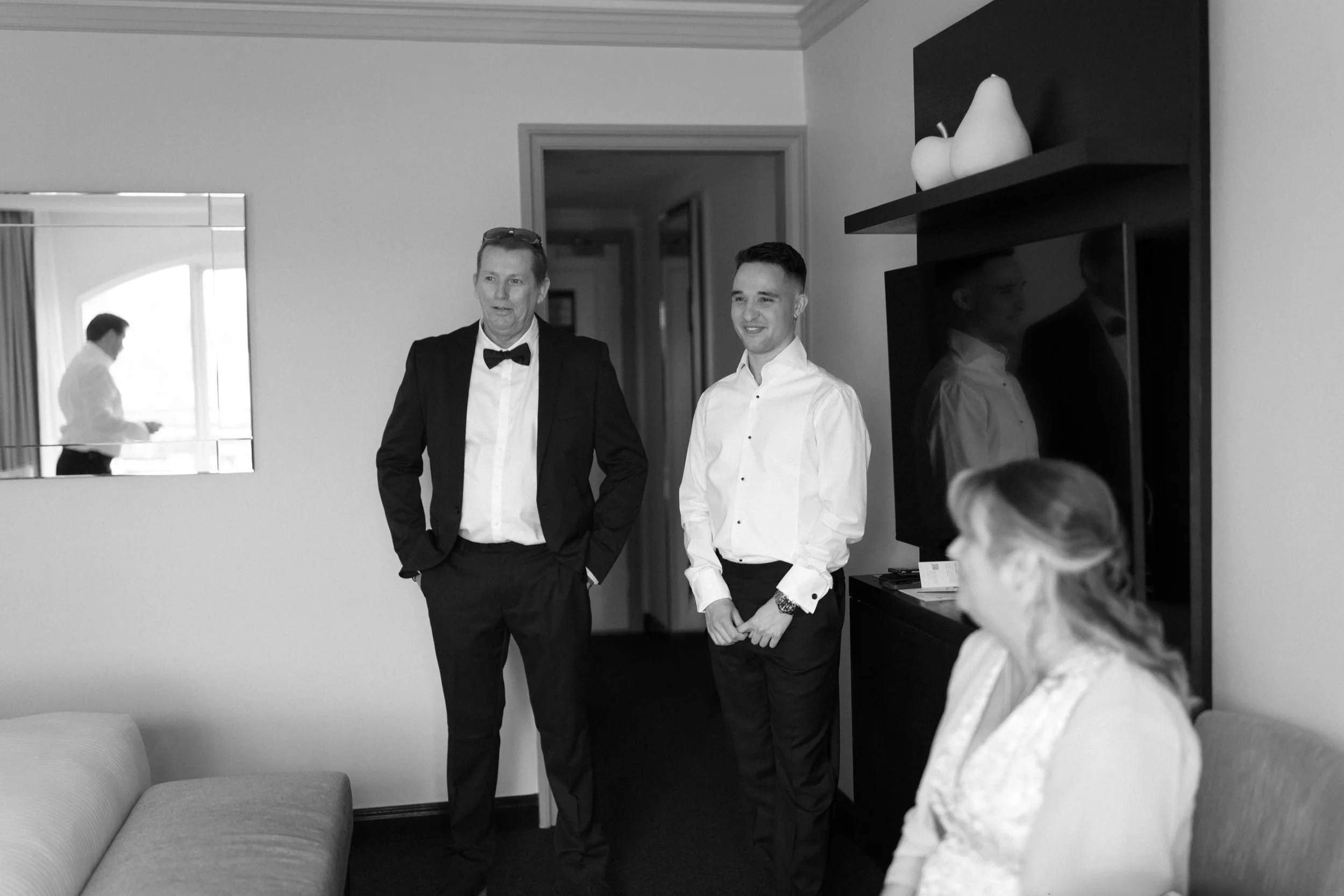 A black and white photo of two men in formal attire standing inside a room, one in a tuxedo with a bow tie and the other in a dress shirt and slacks, with a woman sitting on a couch in the foreground and a man looking at his phone in the background t