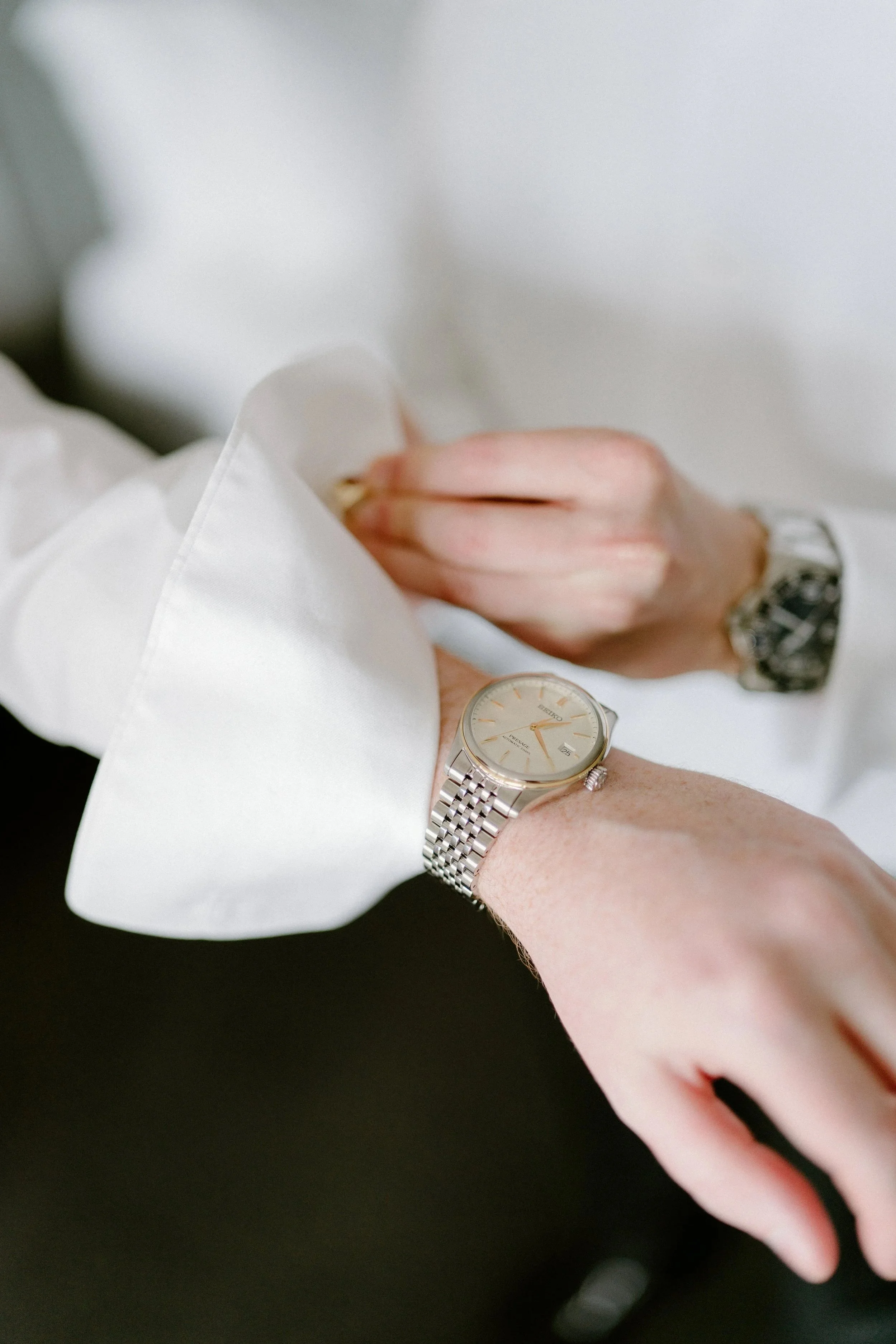 Close-up of a person's wrist showing a silver Seiko watch with a cream face and gold accents, wearing a white long-sleeve shirt.