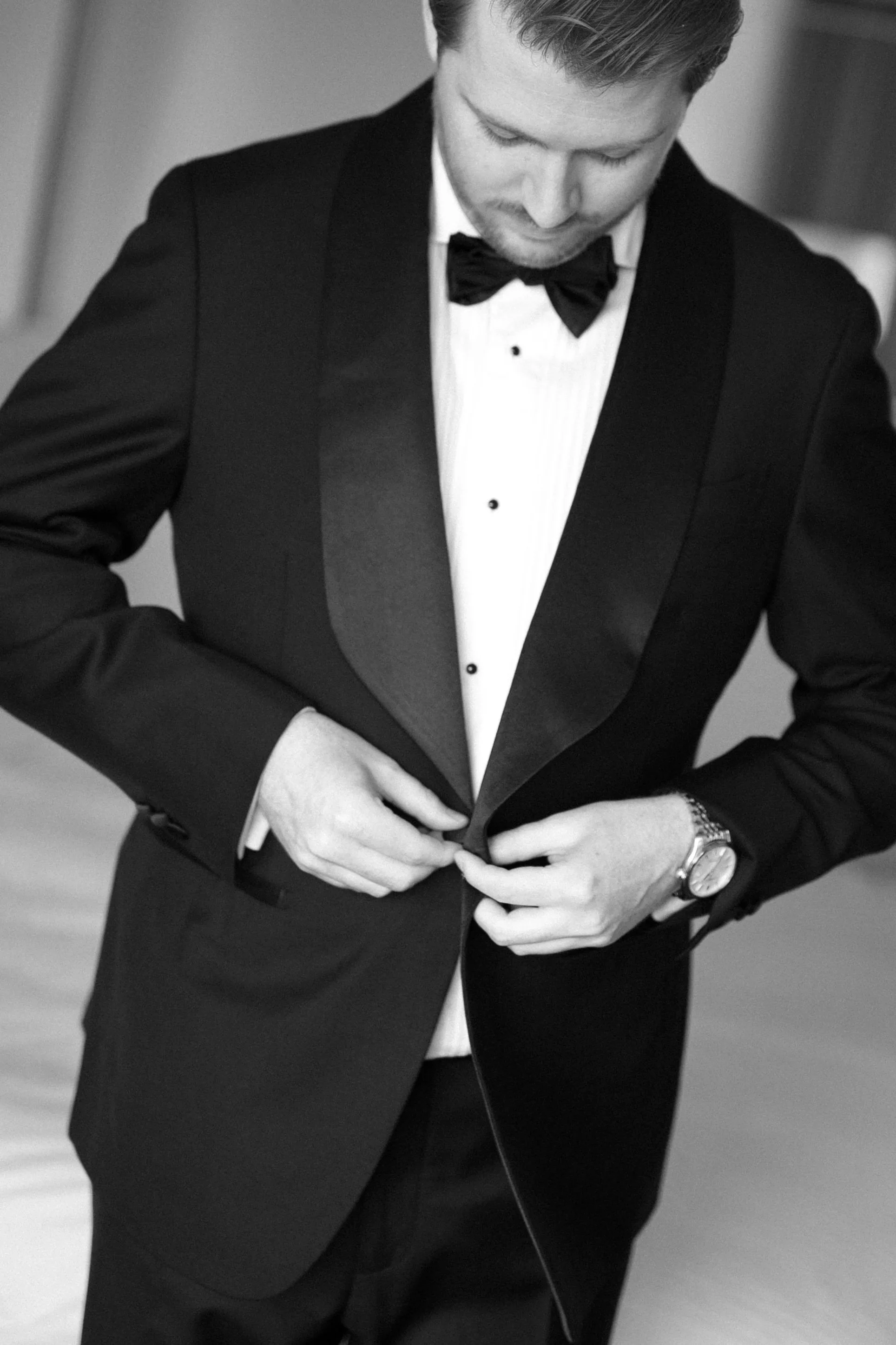 A man in a black tuxedo with a bow tie is buttoning his jacket, looking down. He is wearing a watch on his left wrist.