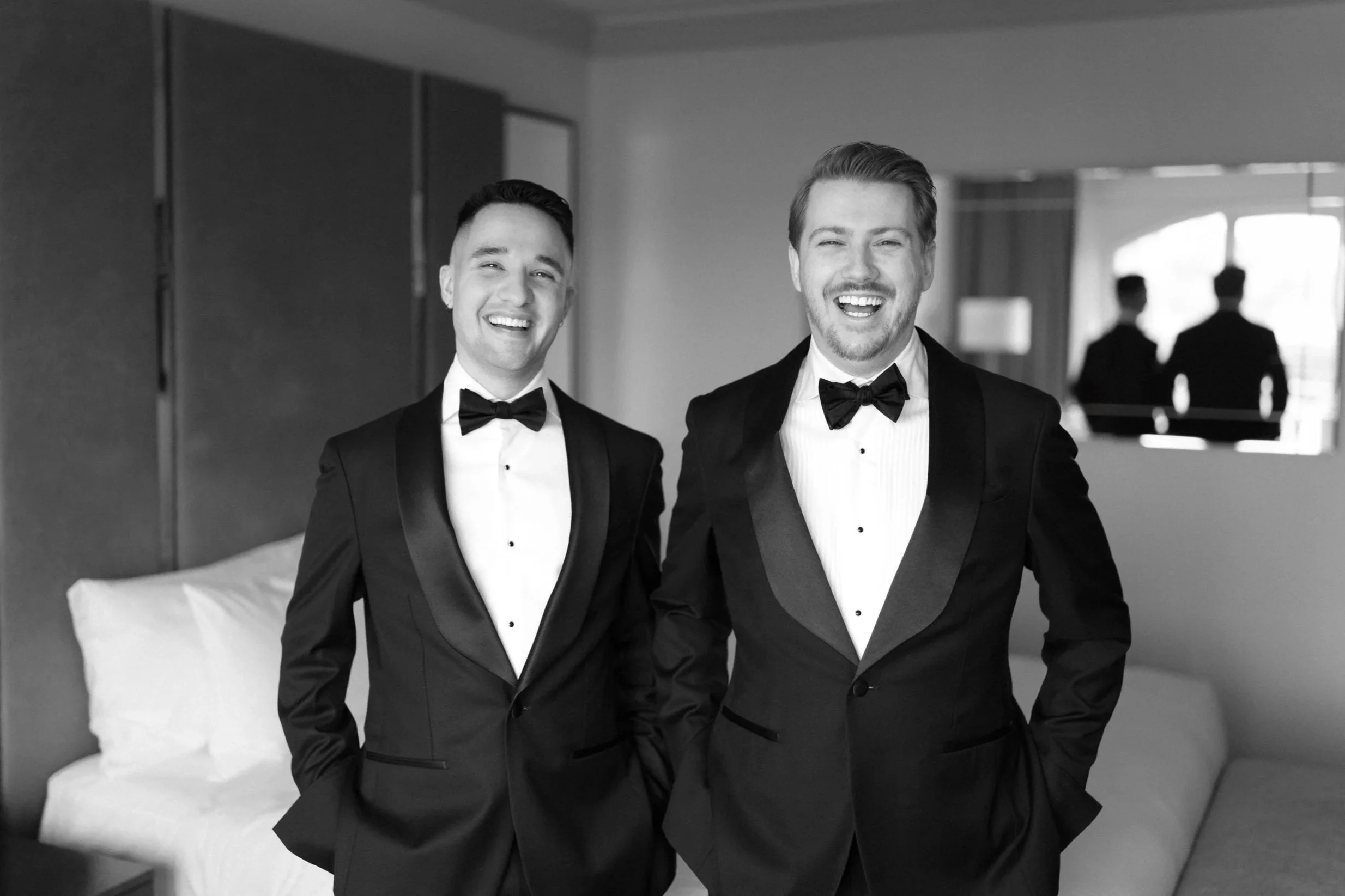 Two men in tuxedos smiling and laughing, standing in a hotel room with a bed and mirror.
