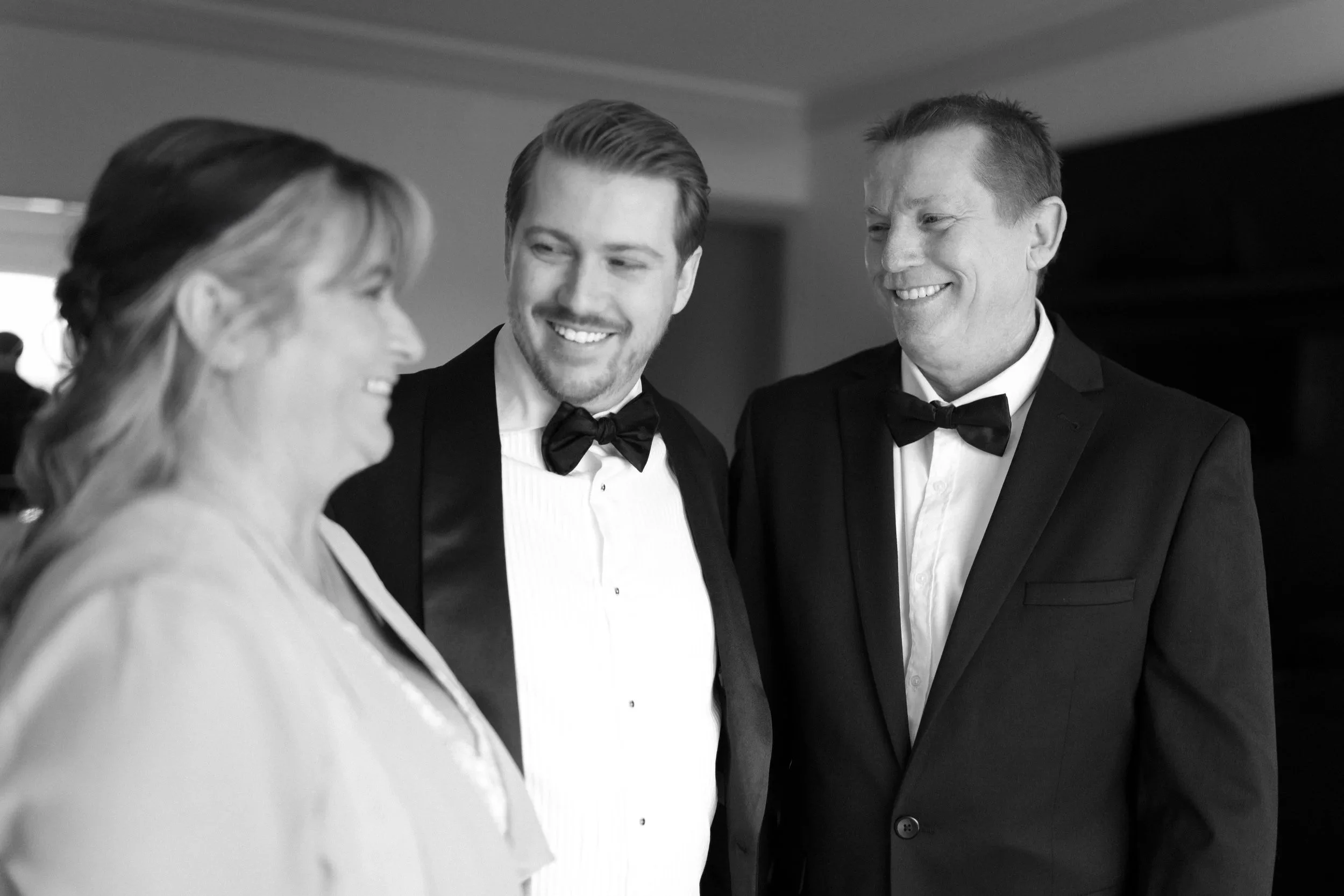 Three people dressed in formal tuxedos and gowns smiling and talking indoors.