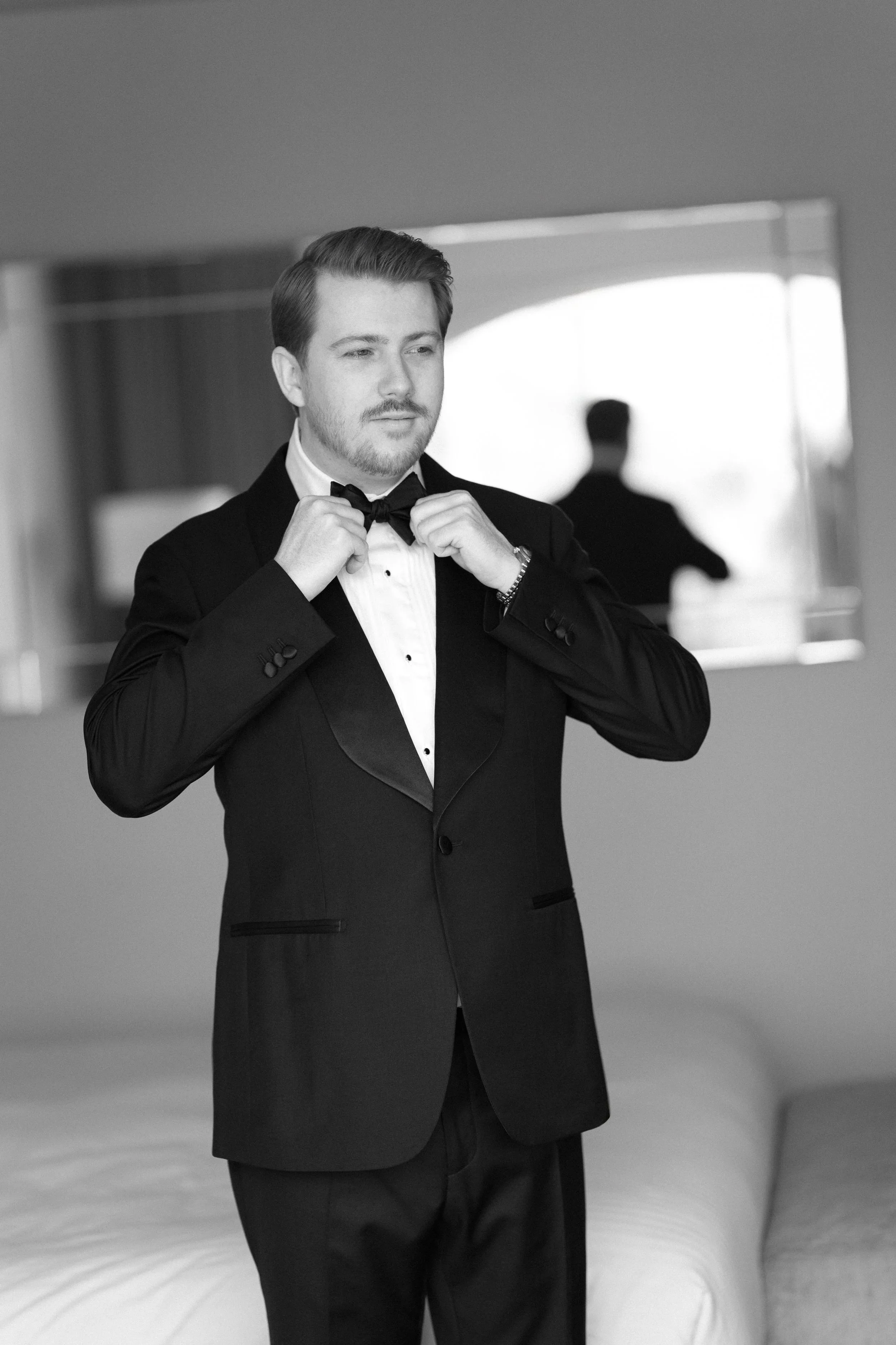 A man in formal tuxedo adjusting his bow tie in front of a mirror.