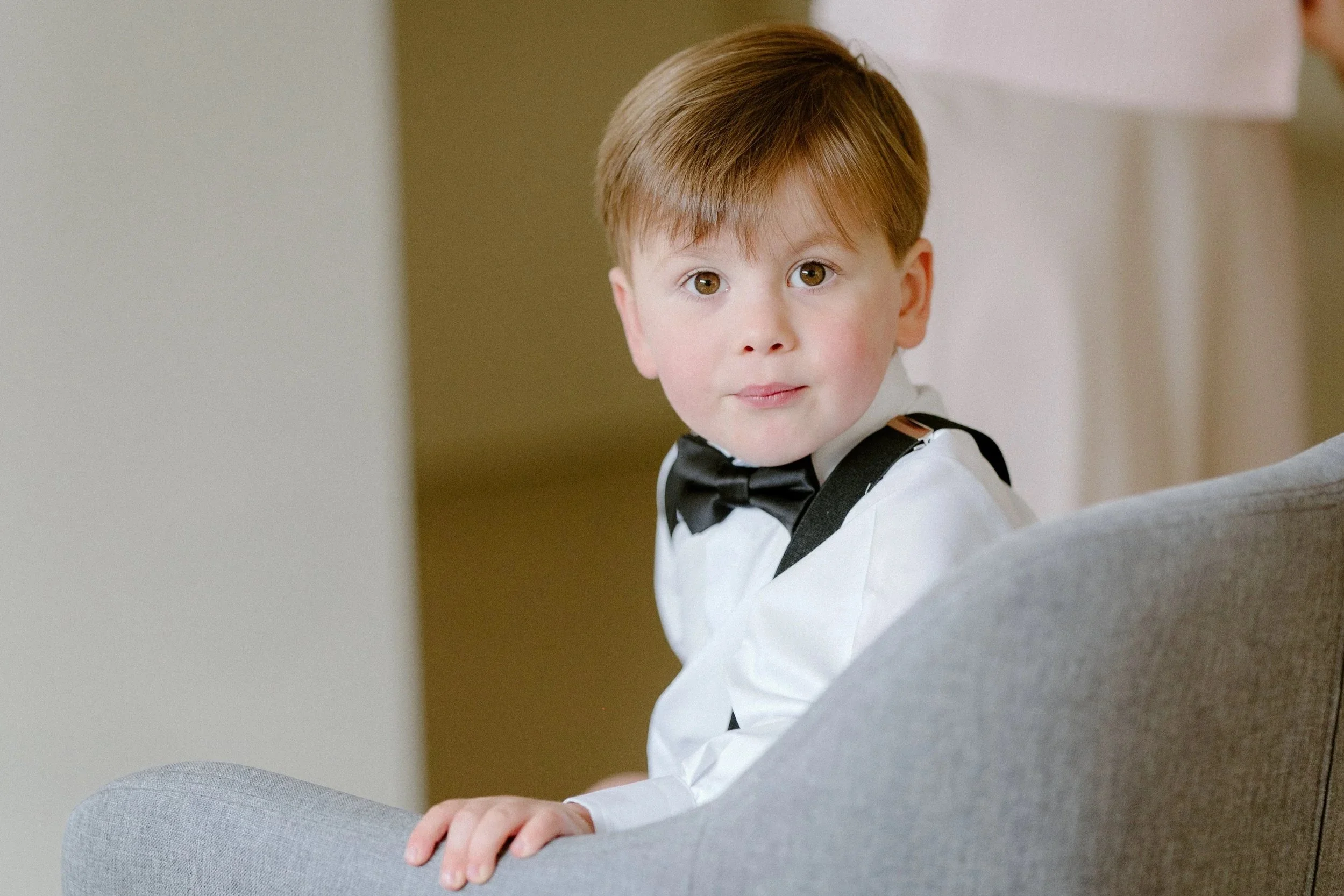 A young boy with red hair and brown eyes dressed in a white shirt and black bow tie, sitting on a gray armchair, looking at the camera with a slight smile.