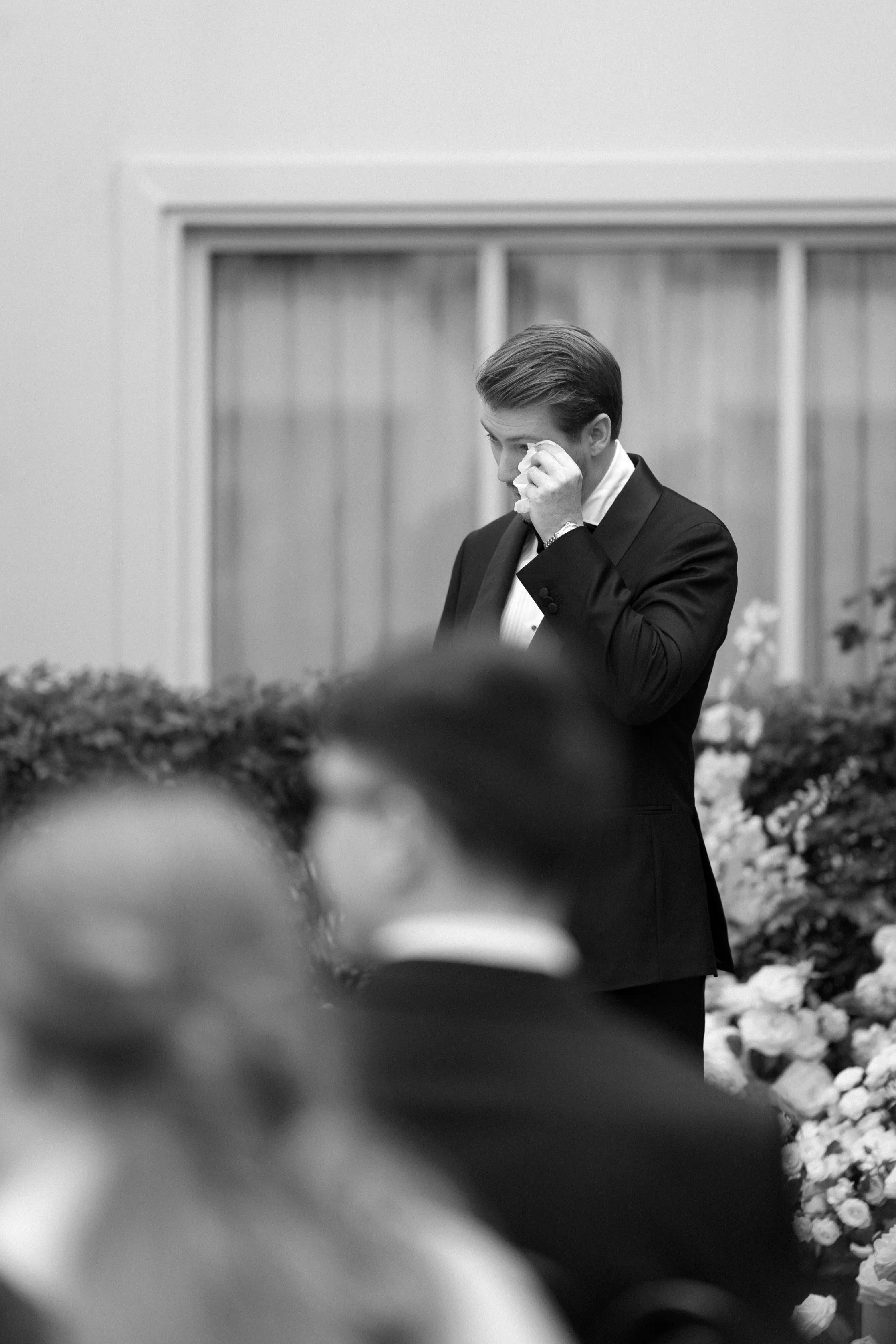 Black and white photo of a man in a tuxedo wiping his eye with a tissue, standing near a floral arrangement, during a formal event.