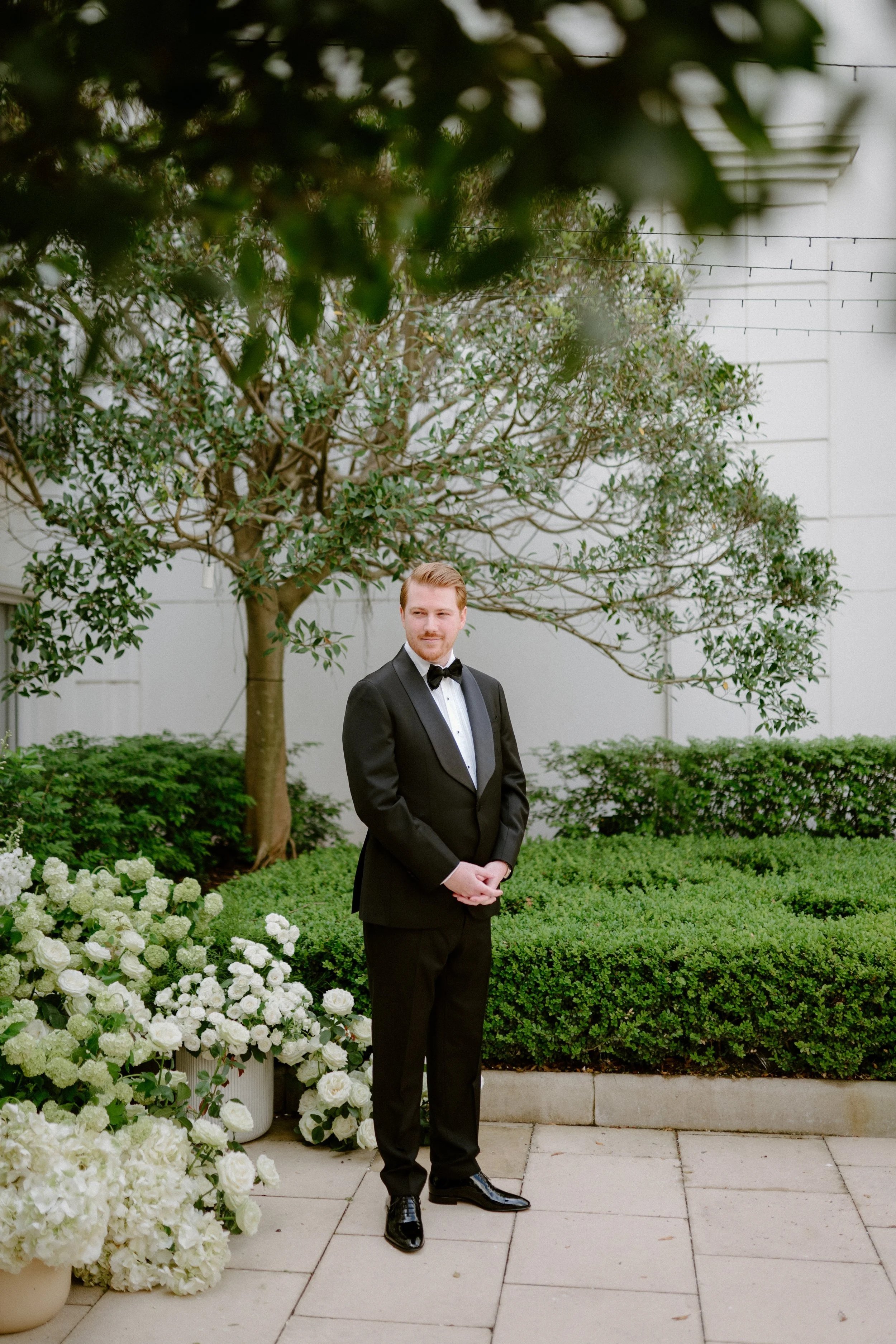 A man in a black tuxedo with a bow tie, standing outdoors next to potted white flowers, with a tree and greenery in the background.