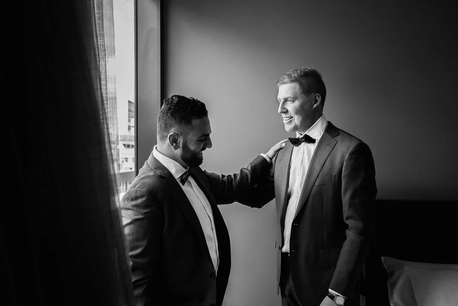 Two men in tuxedos smiling and talking to each other in a room with natural light coming from a window.