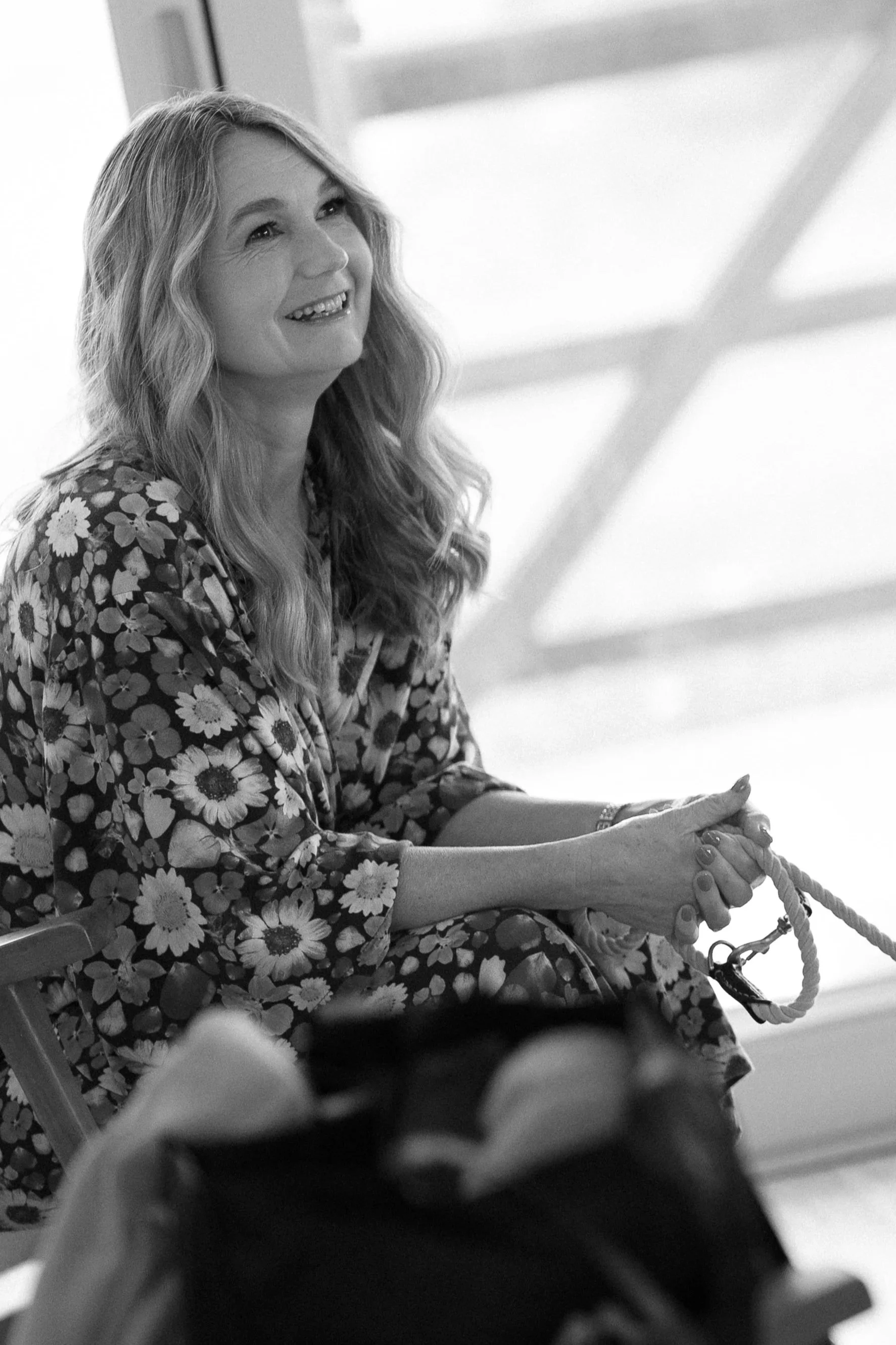 A woman with long wavy hair smiling and sitting by a large window, holding a rope tethered to a dog bed in a grayscale photo.