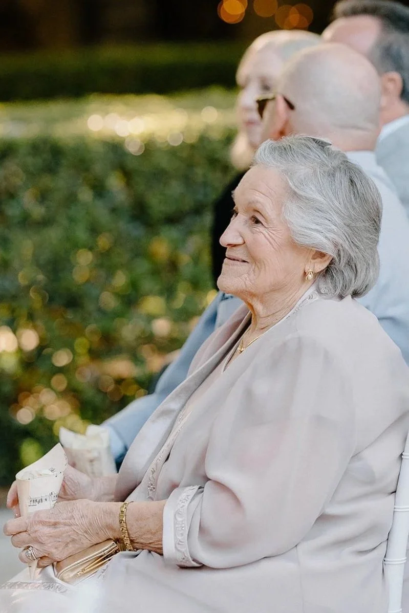 An elderly woman with gray hair sitting outdoors during an event, wearing a beige outfit and holding a cone-shaped item, with blurred people and warm lighting in the background.