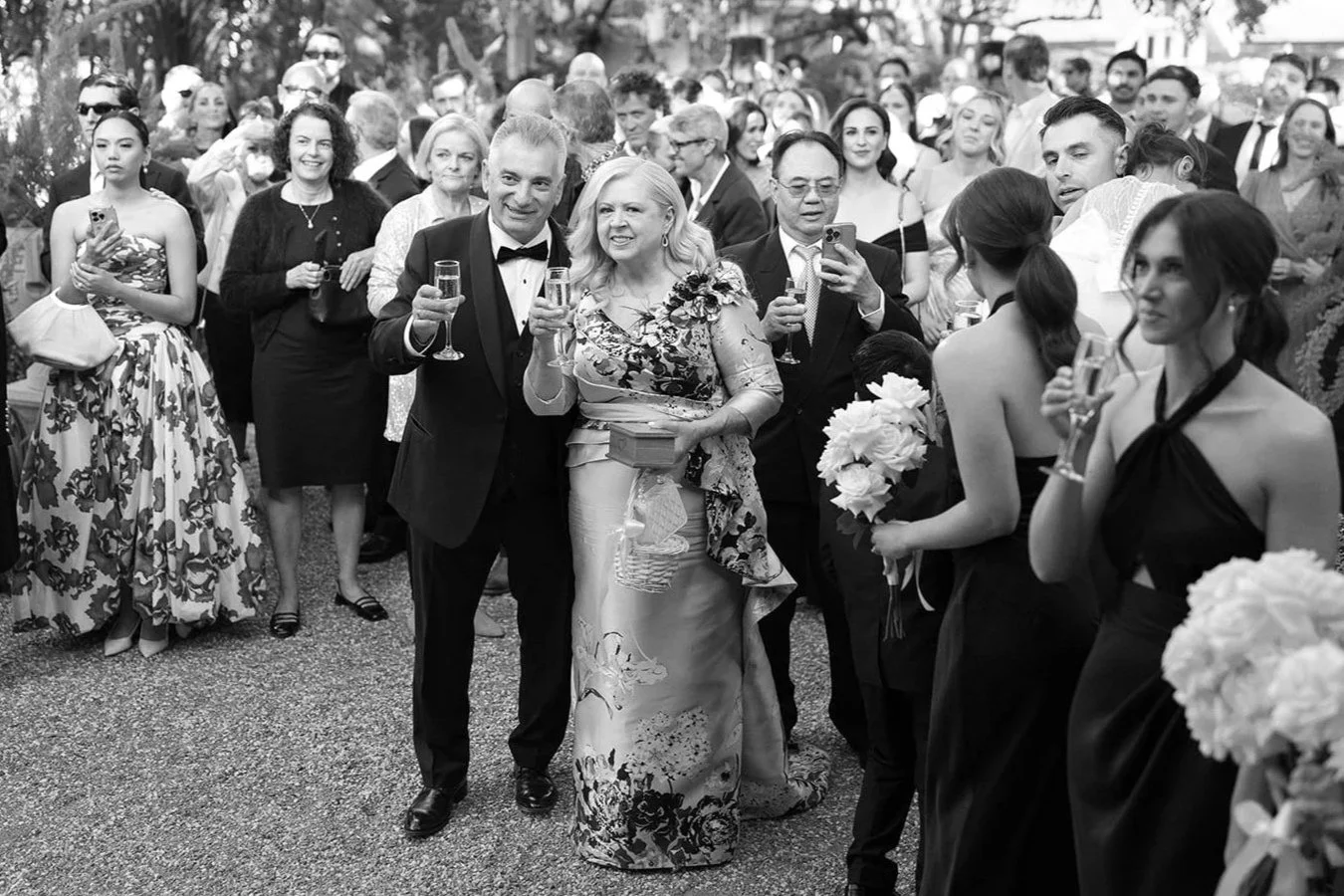 People attending a wedding celebration outdoors, holding glasses of champagne, with some taking photos and others smiling, surrounded by trees and garden setting.