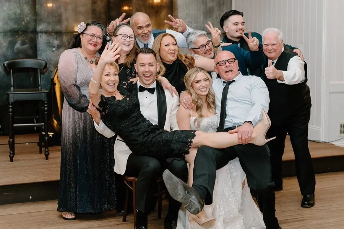 A group of people celebrating at a wedding, with the bride and groom sitting in the center, surrounded by friends and family, all smiling and making playful gestures.