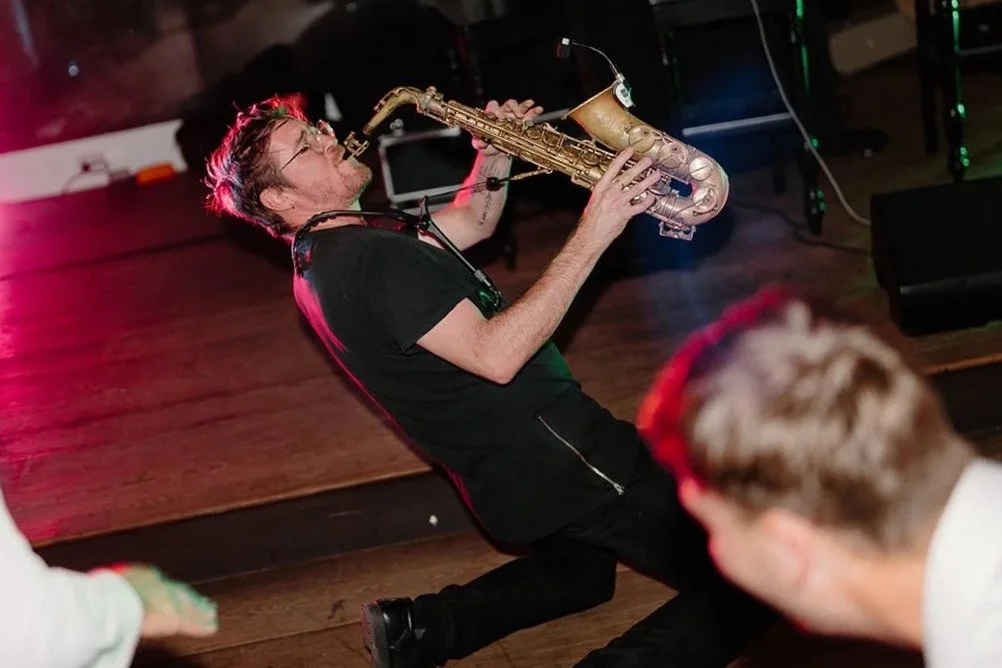 A man playing a saxophone energetically at a party, surrounded by dancing people, with colorful stage lights in the background.