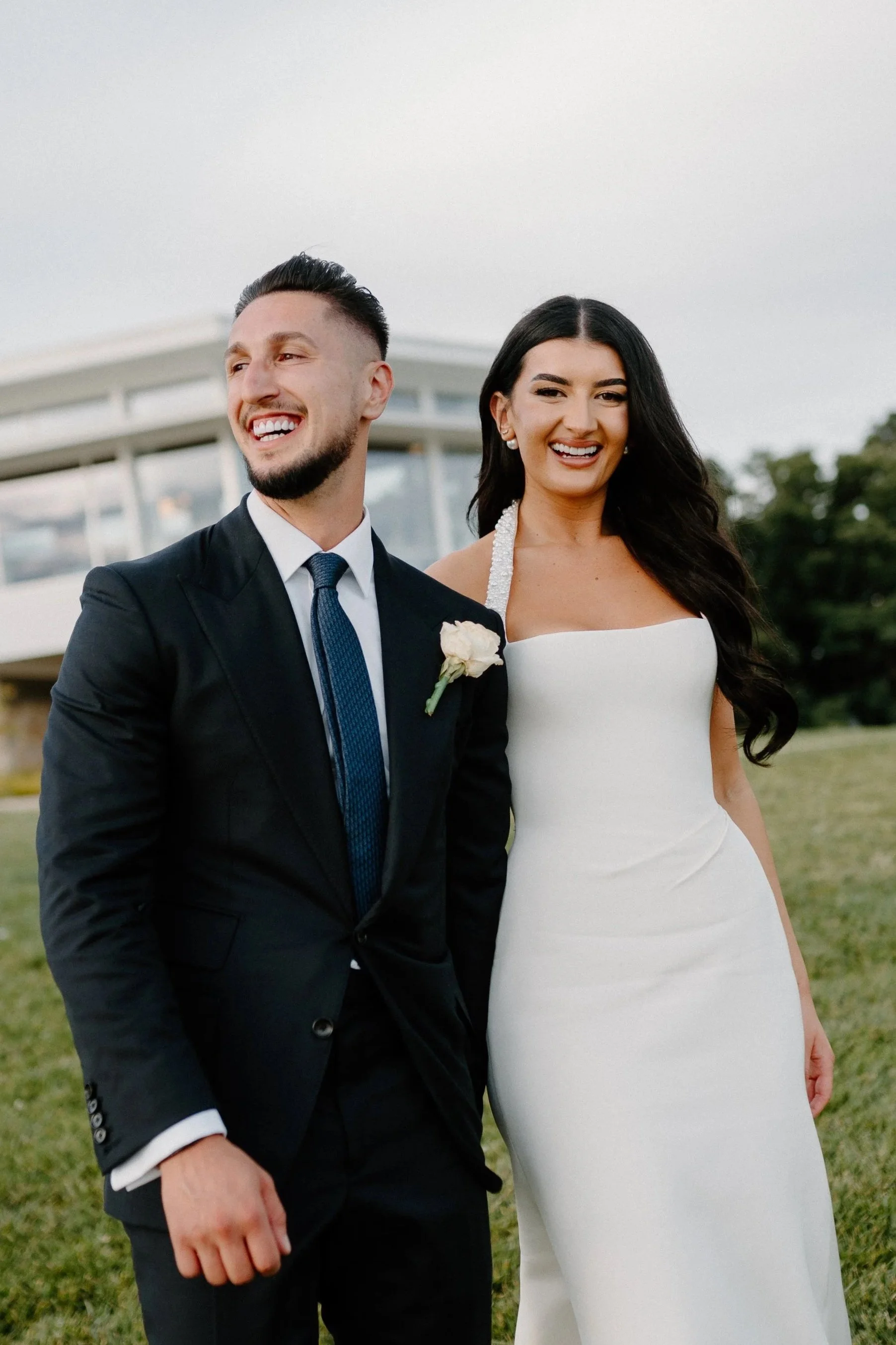 A newlywed couple standing on a grassy field in wedding attire, smiling and enjoying a moment together outdoors, with a modern building in the background.