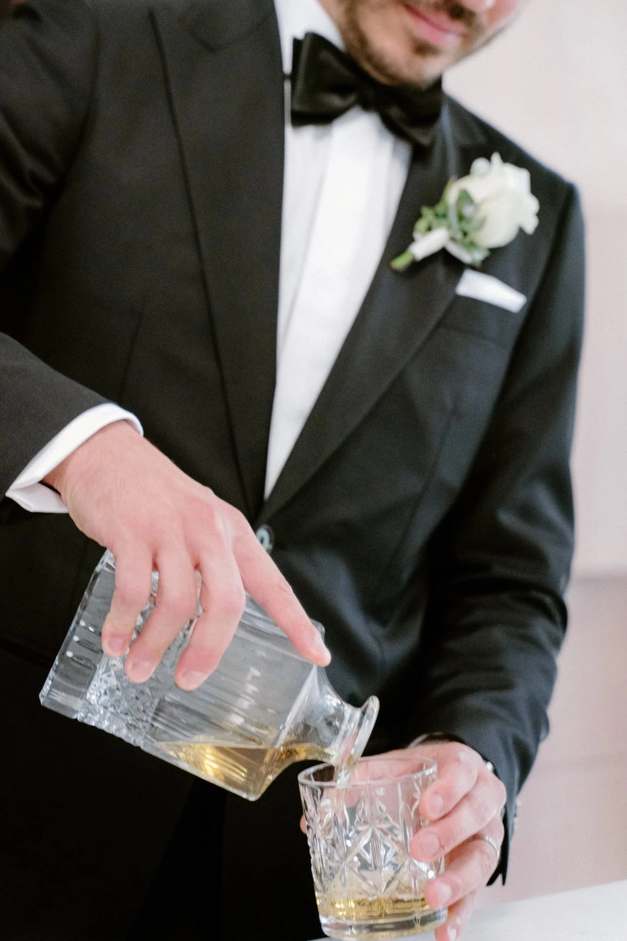 A groom in a tuxedo pouring whiskey into a glass at a wedding reception.