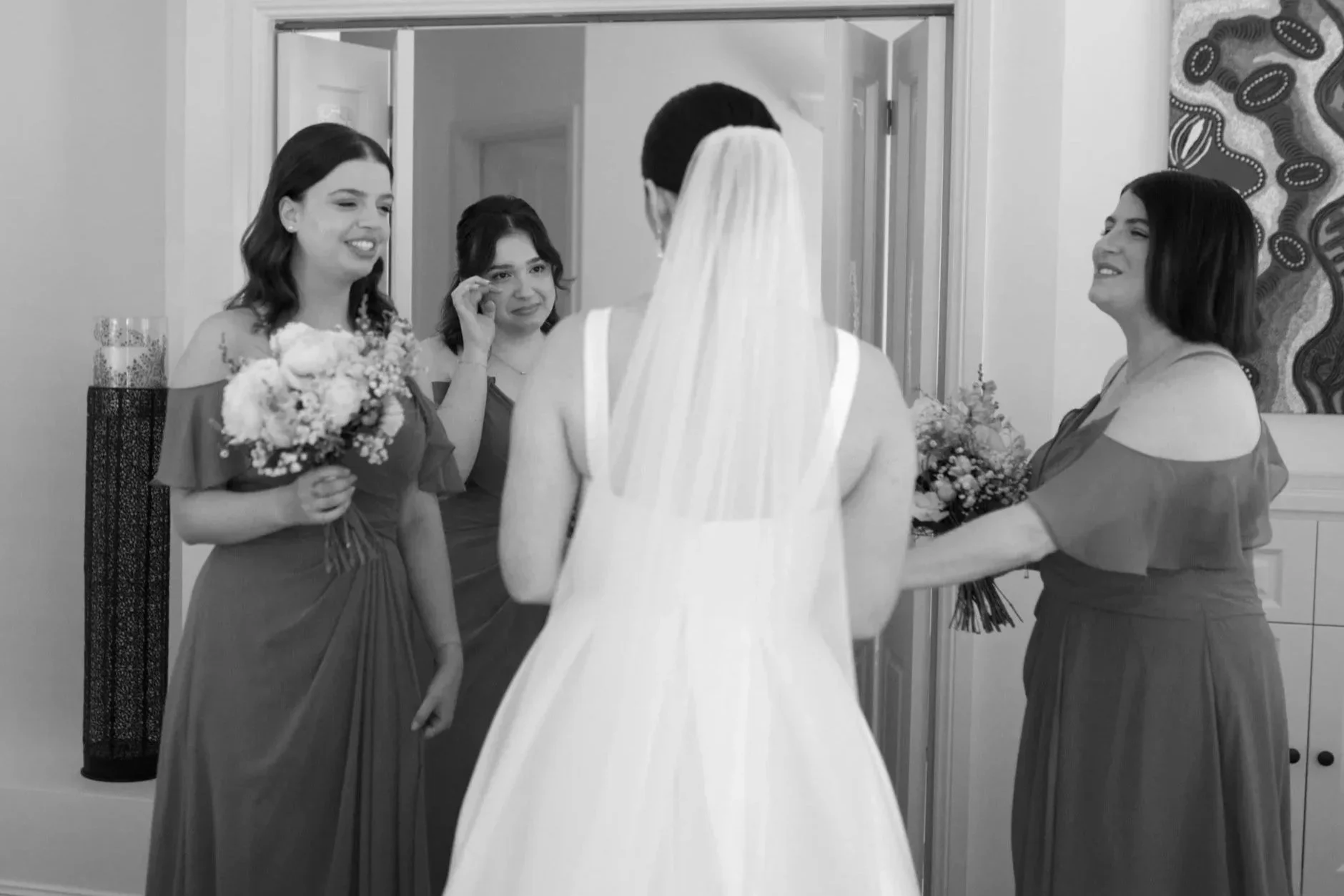 A bride in a white wedding dress and veil is seen from the back, facing four women who are likely her bridesmaids, dressed in matching dresses, with two holding bouquets, inside a room with modern decor and an abstract painting on the wall.