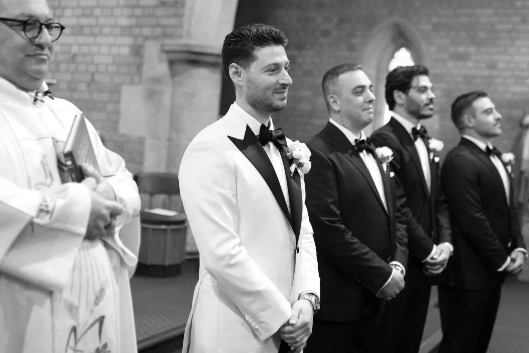 Groom and groomsmen standing in a church during a wedding ceremony, wearing tuxedos with boutonnières, with a priest in the foreground holding a book.