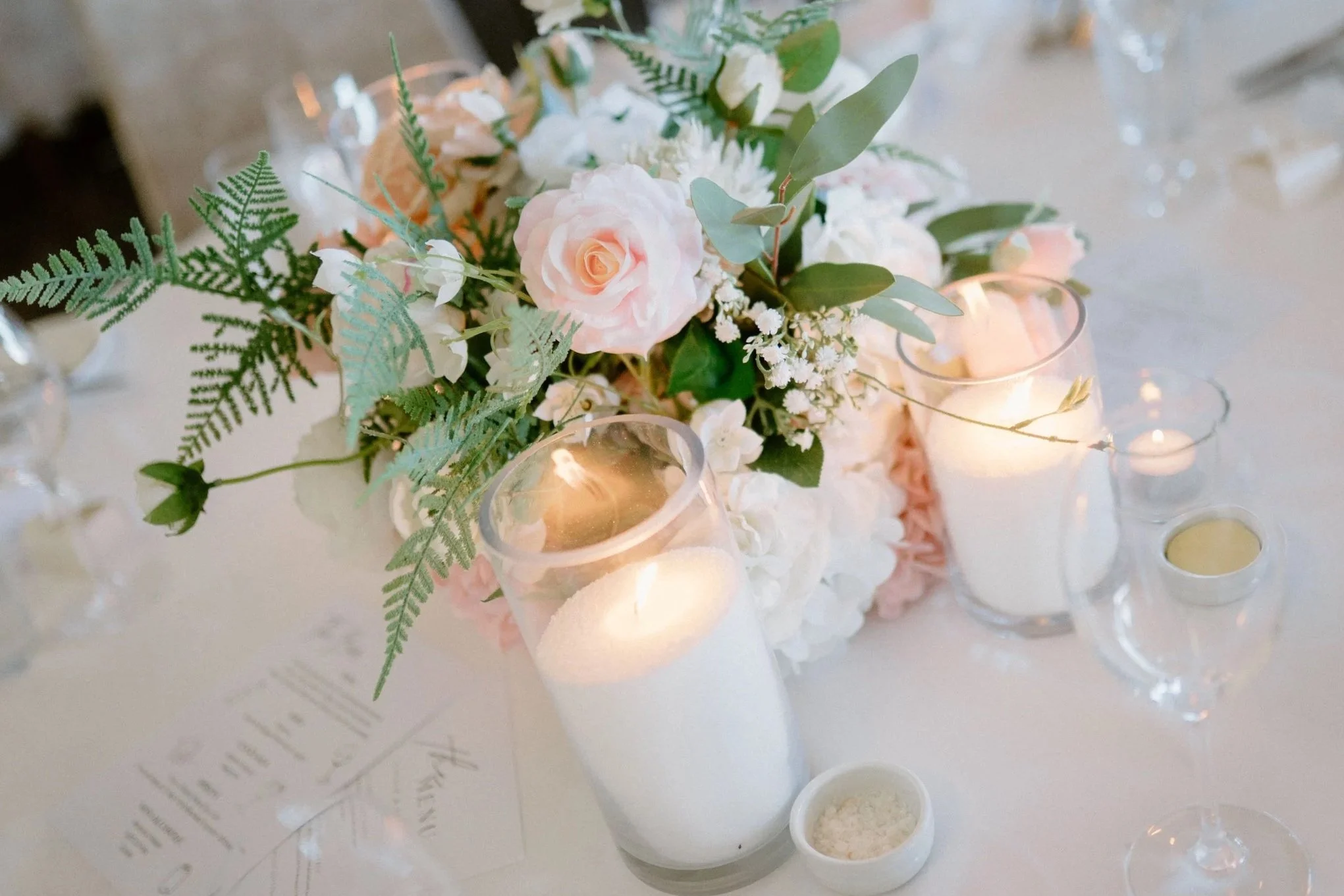 A wedding table centerpiece with pink roses, white flowers, green ferns, and eucalyptus, surrounded by lit candles in glass holders and small bowls, with a menu card and glassware in the background.