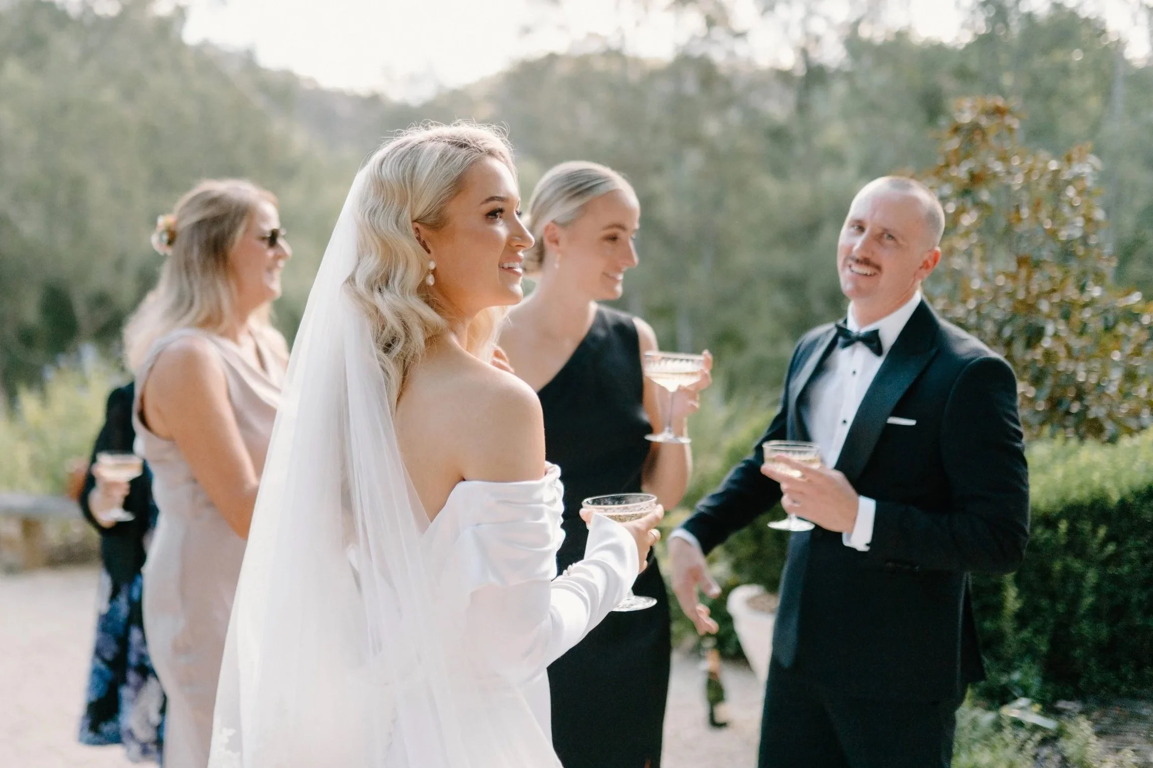 Bride in a white off-the-shoulder gown with long veil and pearl earrings smiling while holding a champagne glass, standing outdoors with three women and a man in tuxedo, celebrating at a wedding reception.