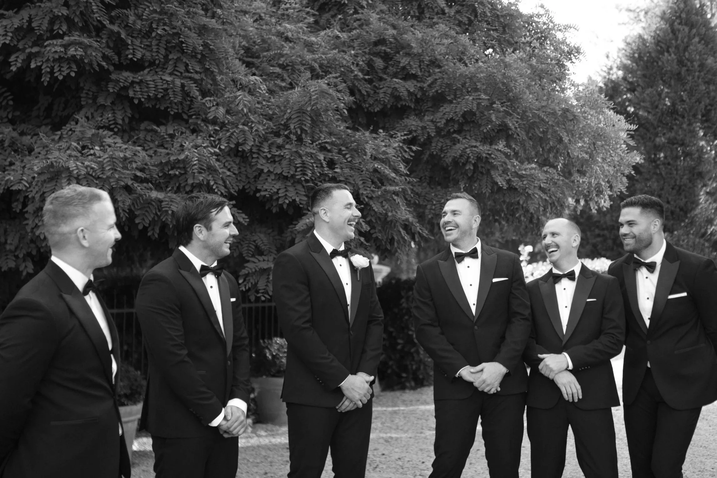 Seven men in tuxedos standing outdoors, laughing and talking, with trees in the background.