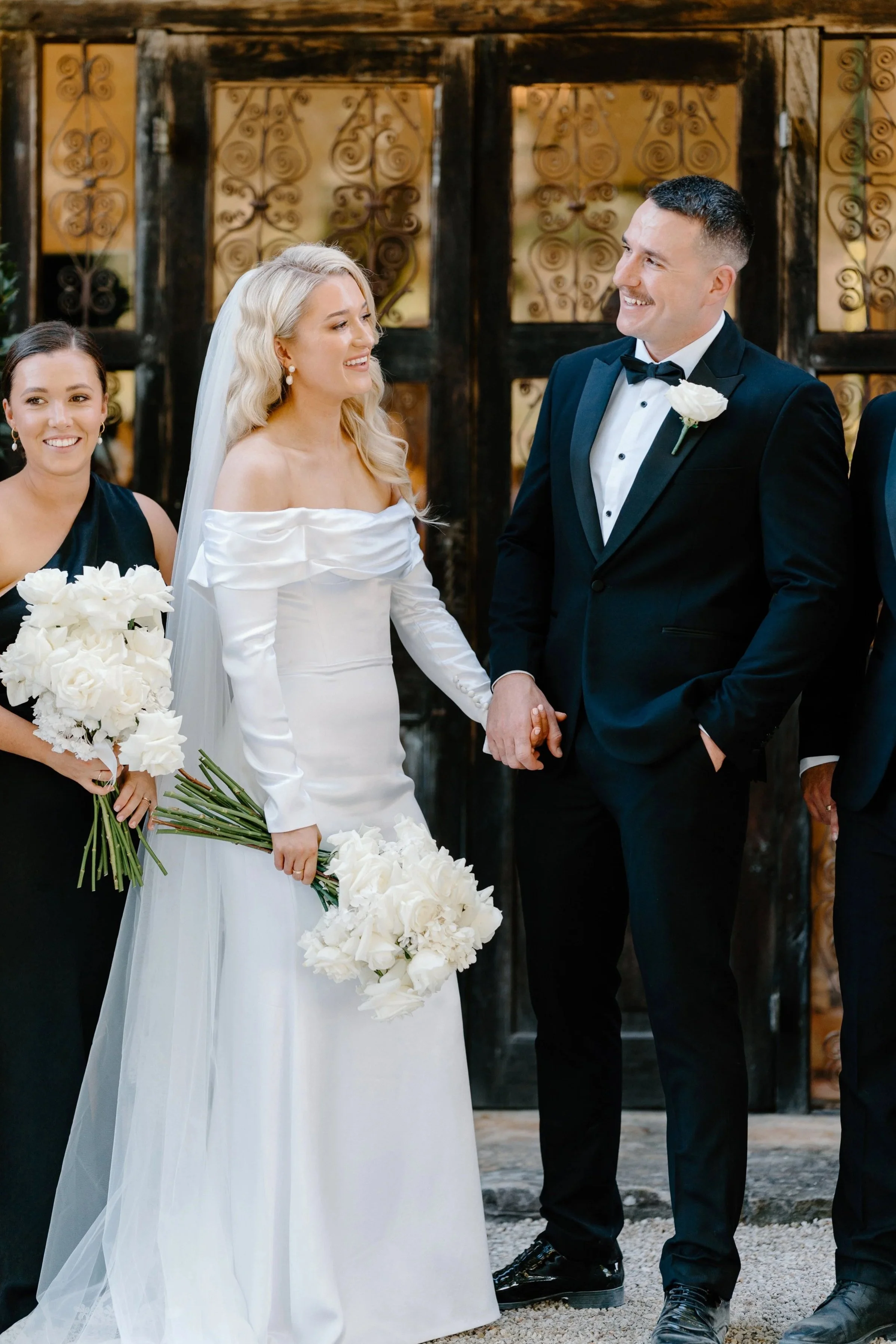 A bride and groom holding hands during their wedding ceremony, with two bridesmaids in the background, all smiling.
