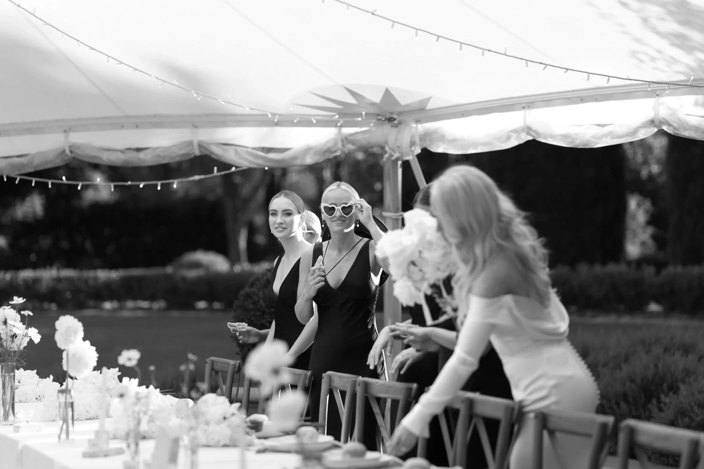 Women celebrating at an outdoor wedding reception under a white canopy, with one woman in a white dress and others in black dresses, surrounded by flowers and decorated tables.