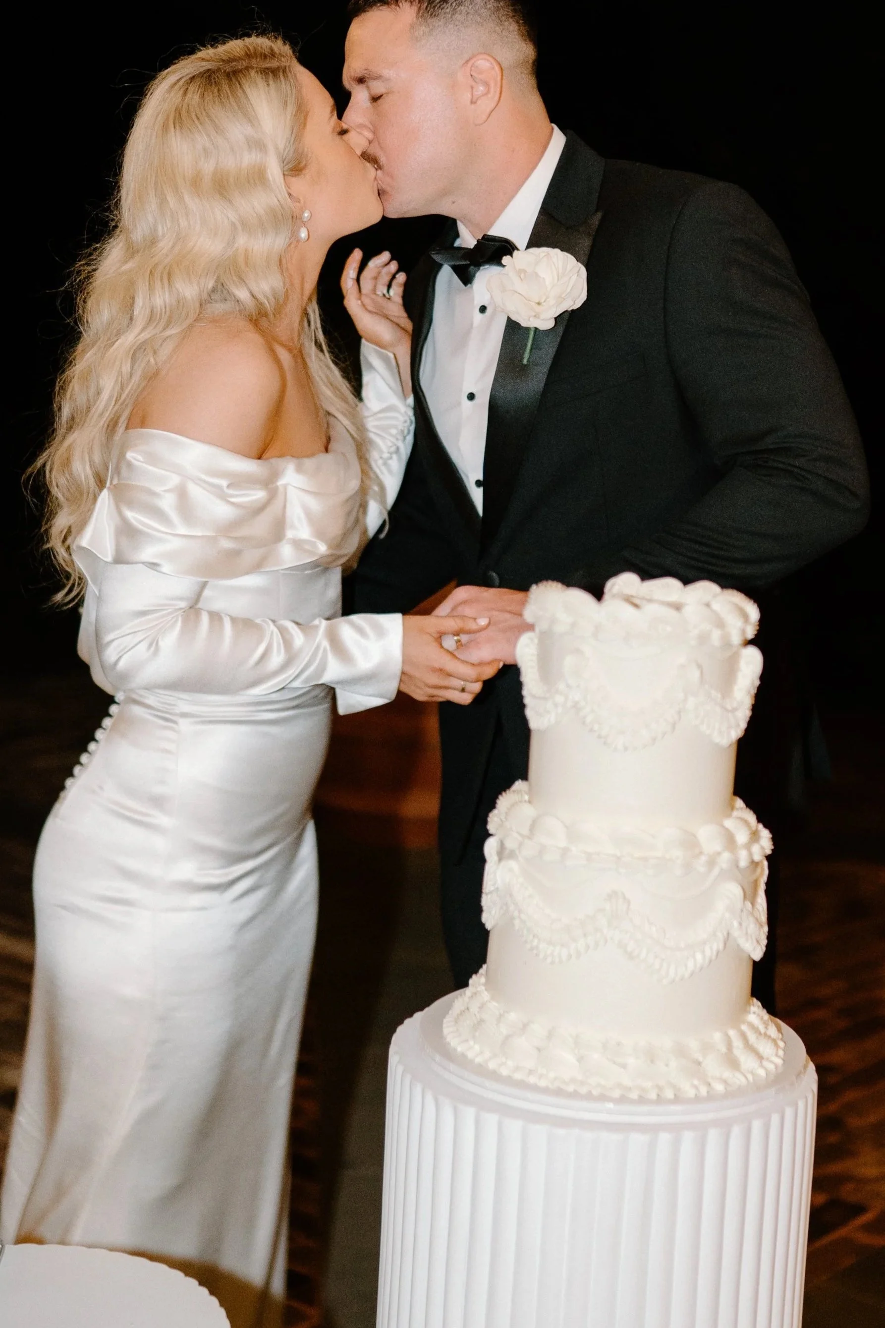 Bride and groom kissing at their wedding, standing behind a large white tiered wedding cake adorned with decorative piping and lace-like details.