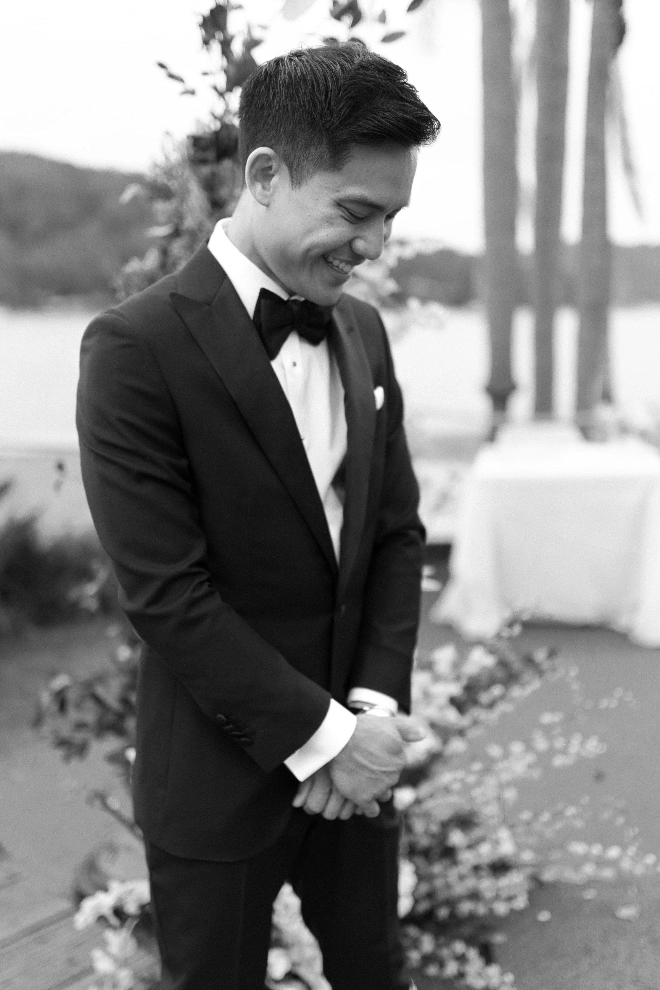 A man in a tuxedo stands outdoors near water, smiling with his head bowed and hands clasped.