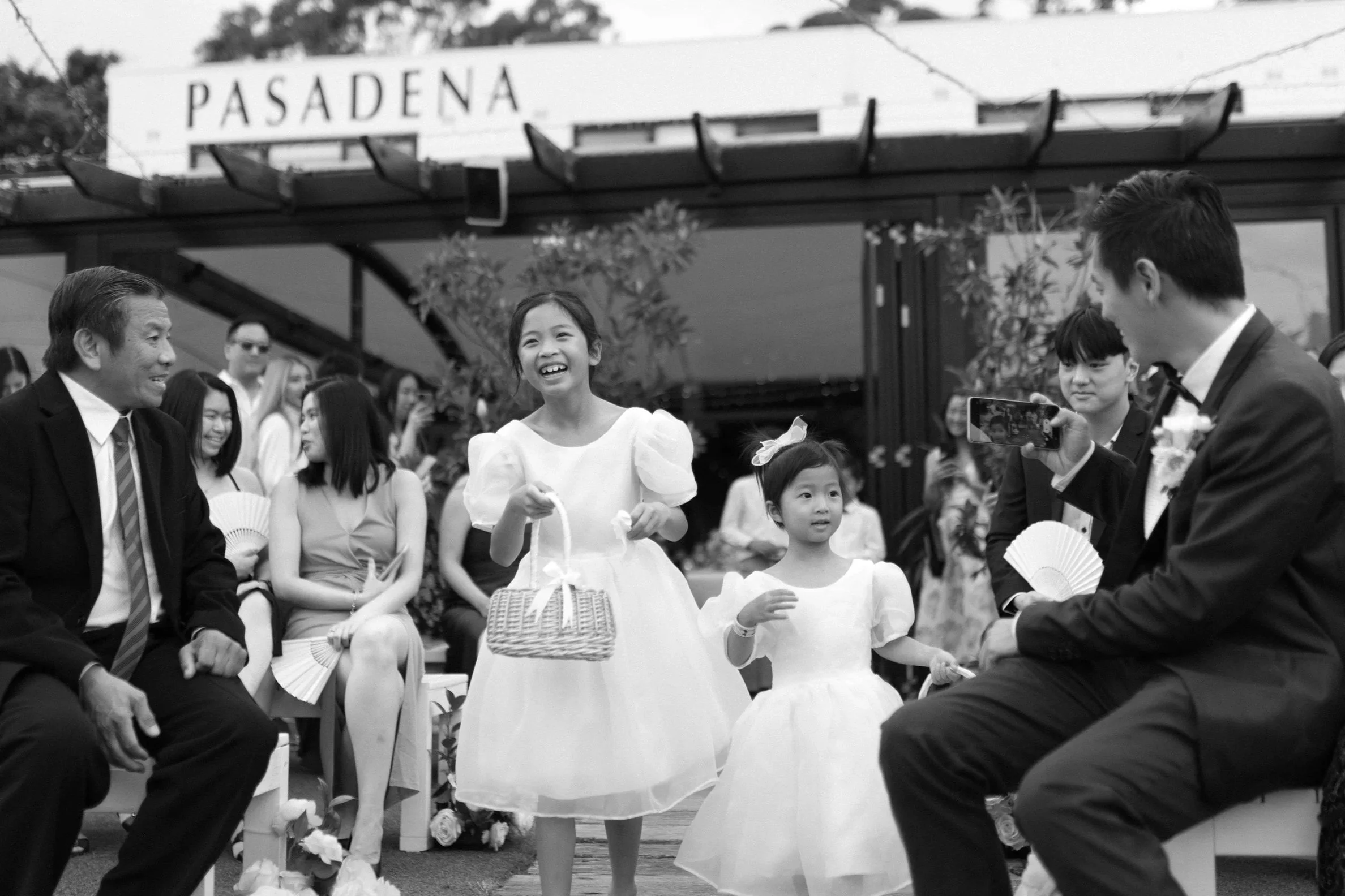 Black and white photo of a wedding ceremony with children and adults smiling, with two young girls in white dresses walking down the aisle, while people seated outdoors watch and record the special moment.