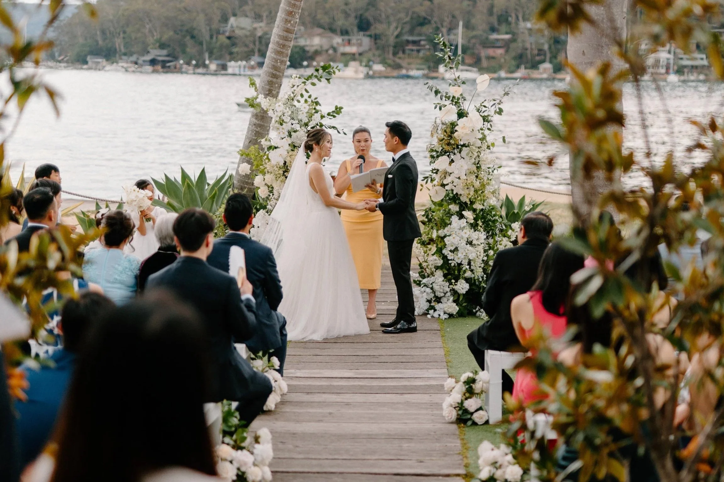 A wedding ceremony takes place outdoors by a lake, with the bride and groom holding hands and exchanging vows. The bride wears a white wedding dress and veil, and the groom wears a black suit. An officiant stands between them, holding a microphone an