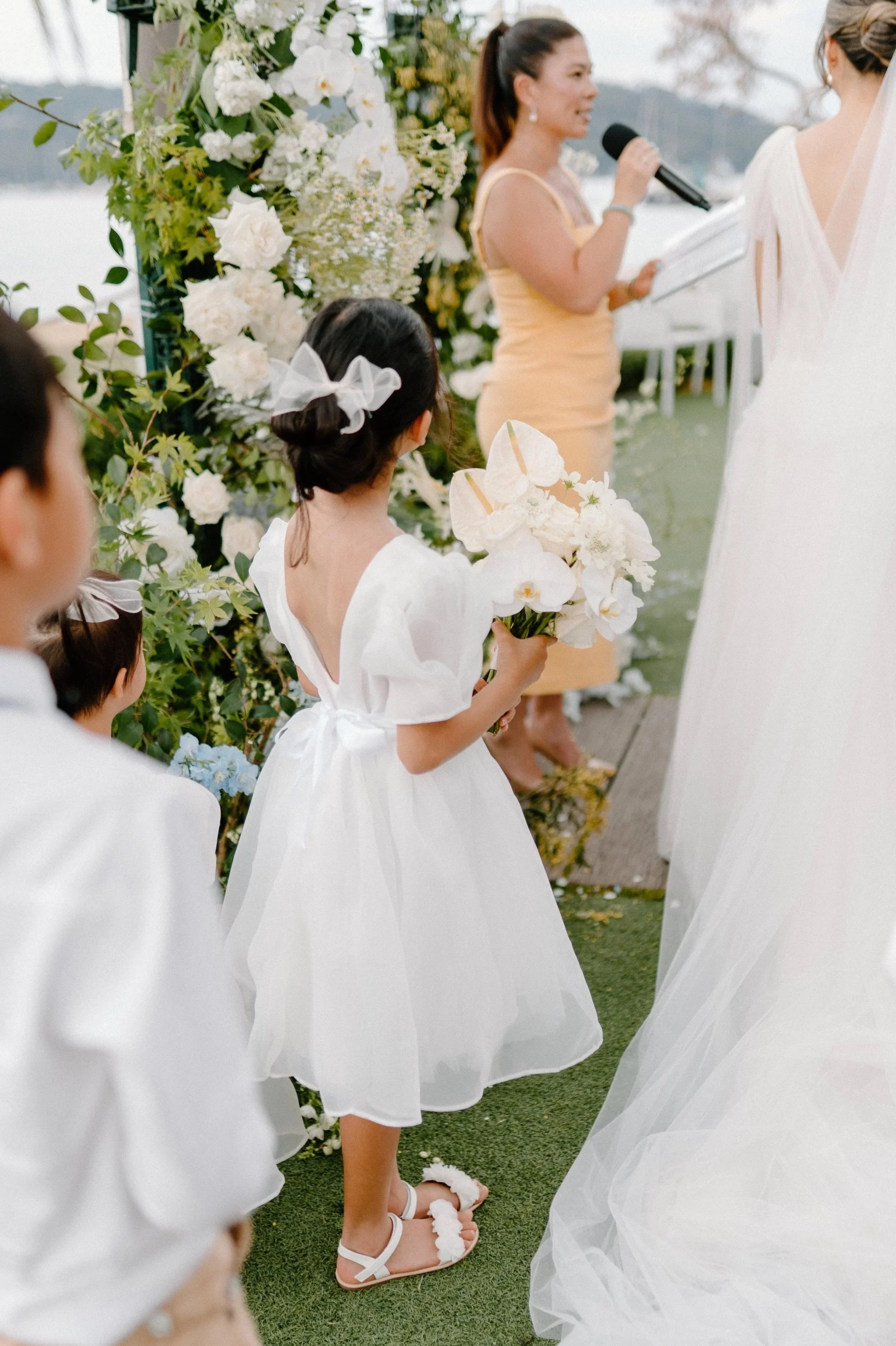 A wedding ceremony outdoors with a bride in a white gown and veil, a woman officiant speaking into a microphone, and children in white dresses holding flowers, all by a floral arch near a body of water.