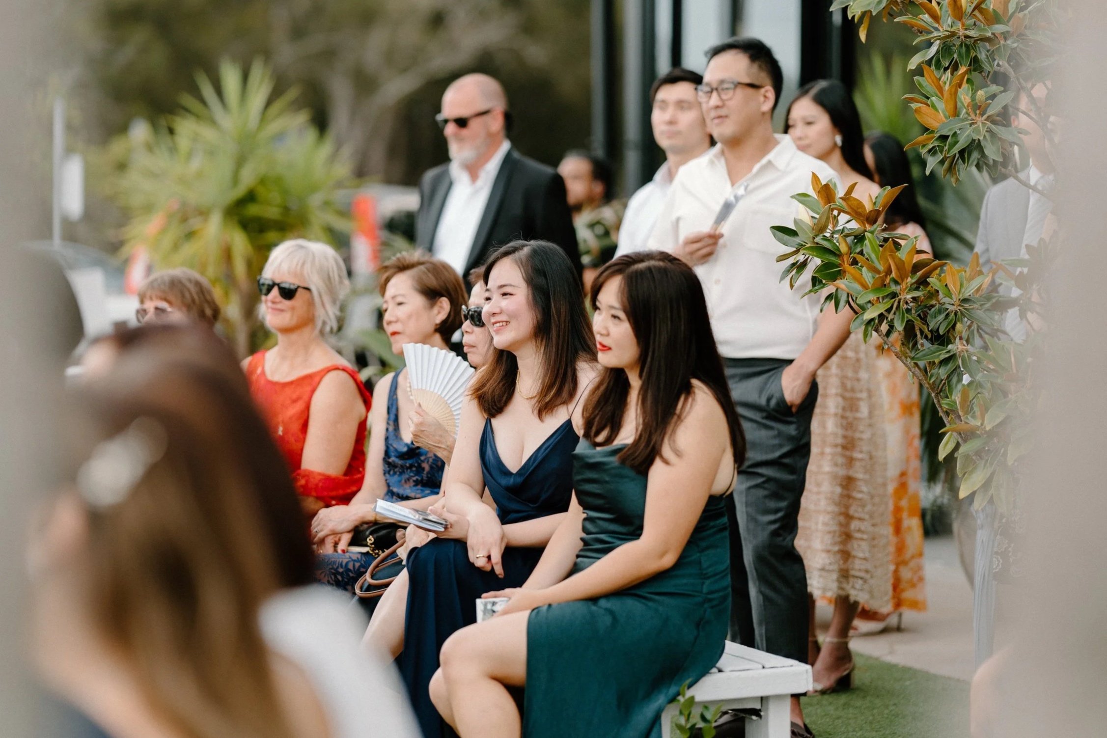 Group of people gathered outdoors, some sitting and some standing, watching an event with greenery and trees in the background. The seated women are dressed in elegant attire, with one holding a fan, smiling and looking ahead. The standing individual
