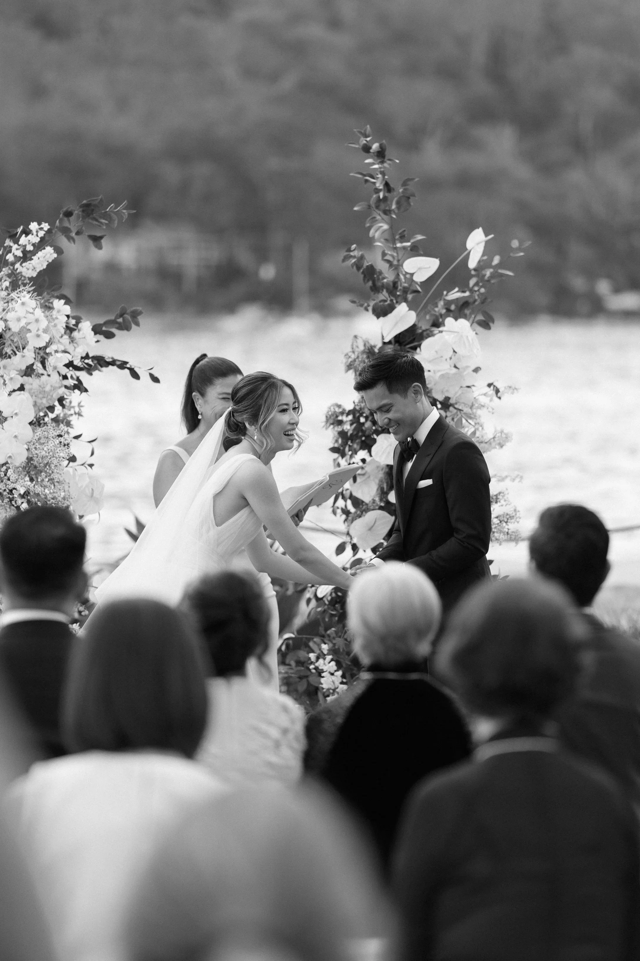 A black and white photo of a wedding ceremony outdoors by the water with arch decorated with flowers, where a bride and groom are exchanging vows.