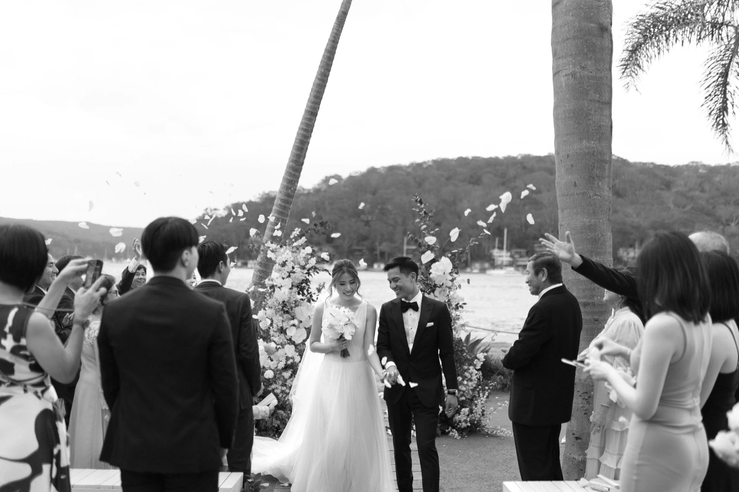 A wedding ceremony outdoors by the water, with a bride and groom holding hands and smiling, surrounded by guests, trees, and mountain scenery.