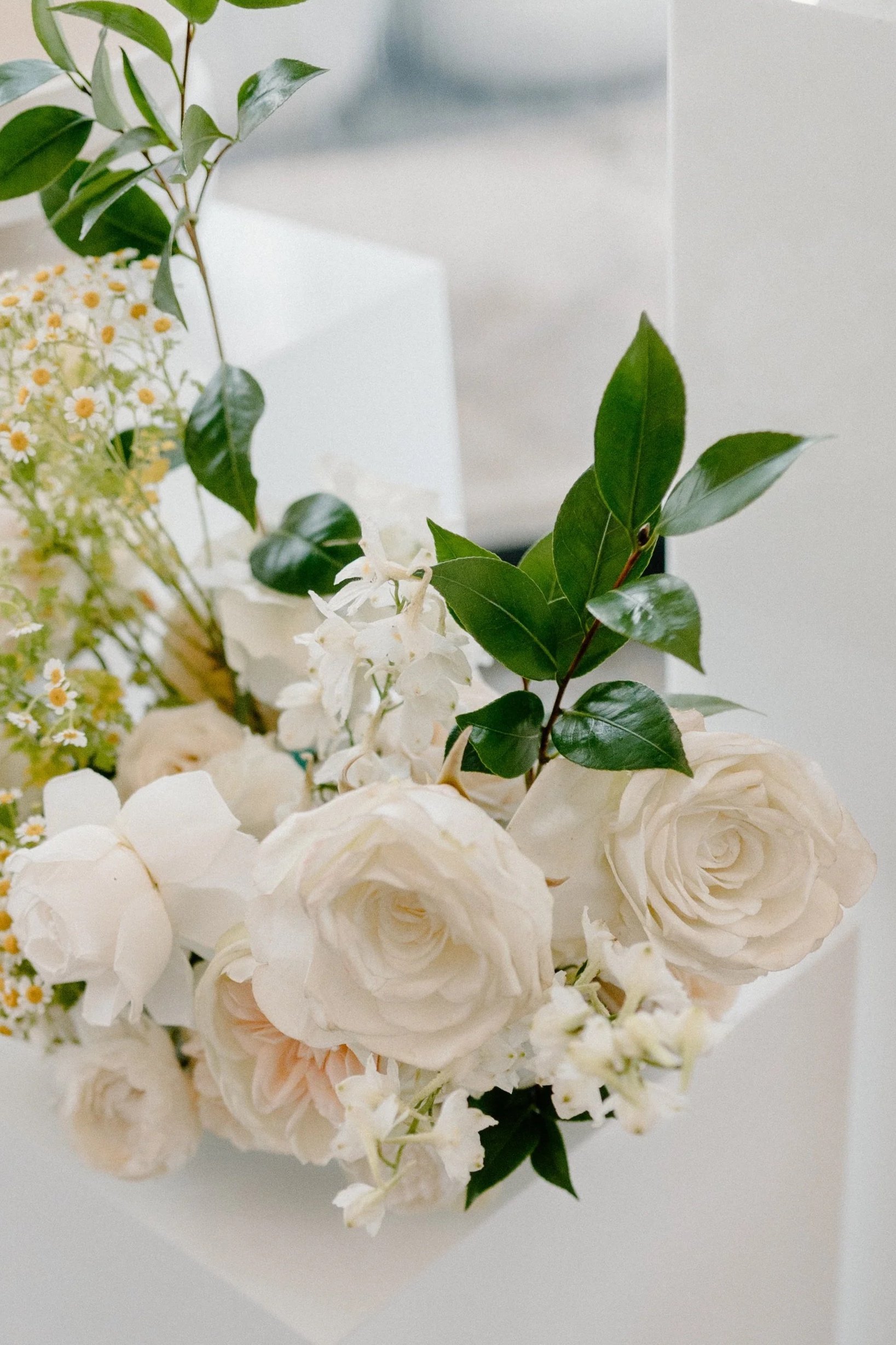 White floral arrangement with roses and greenery in a white rectangular vase, with a candle and decorative items in the background.