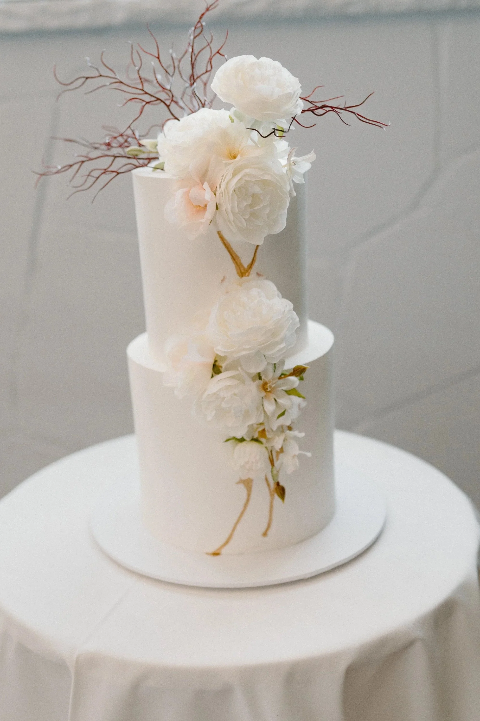 A white two-tiered wedding cake decorated with white flowers and brown curly branches, placed on a round white tablecloth and a white cake board.