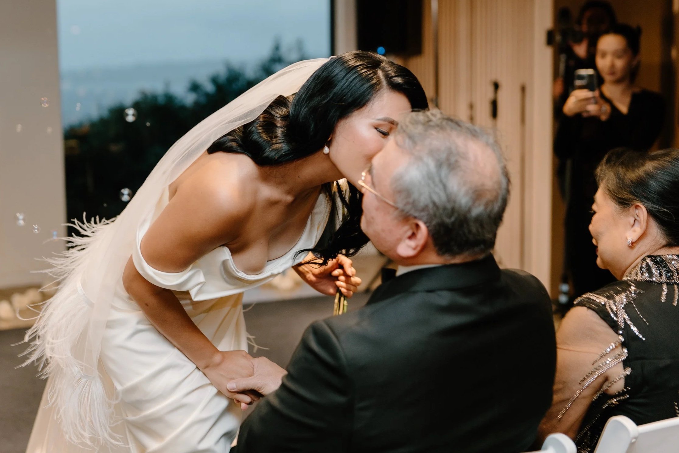 A bride in a white dress and veil leans forward to kiss an older man and woman seated at a wedding reception.