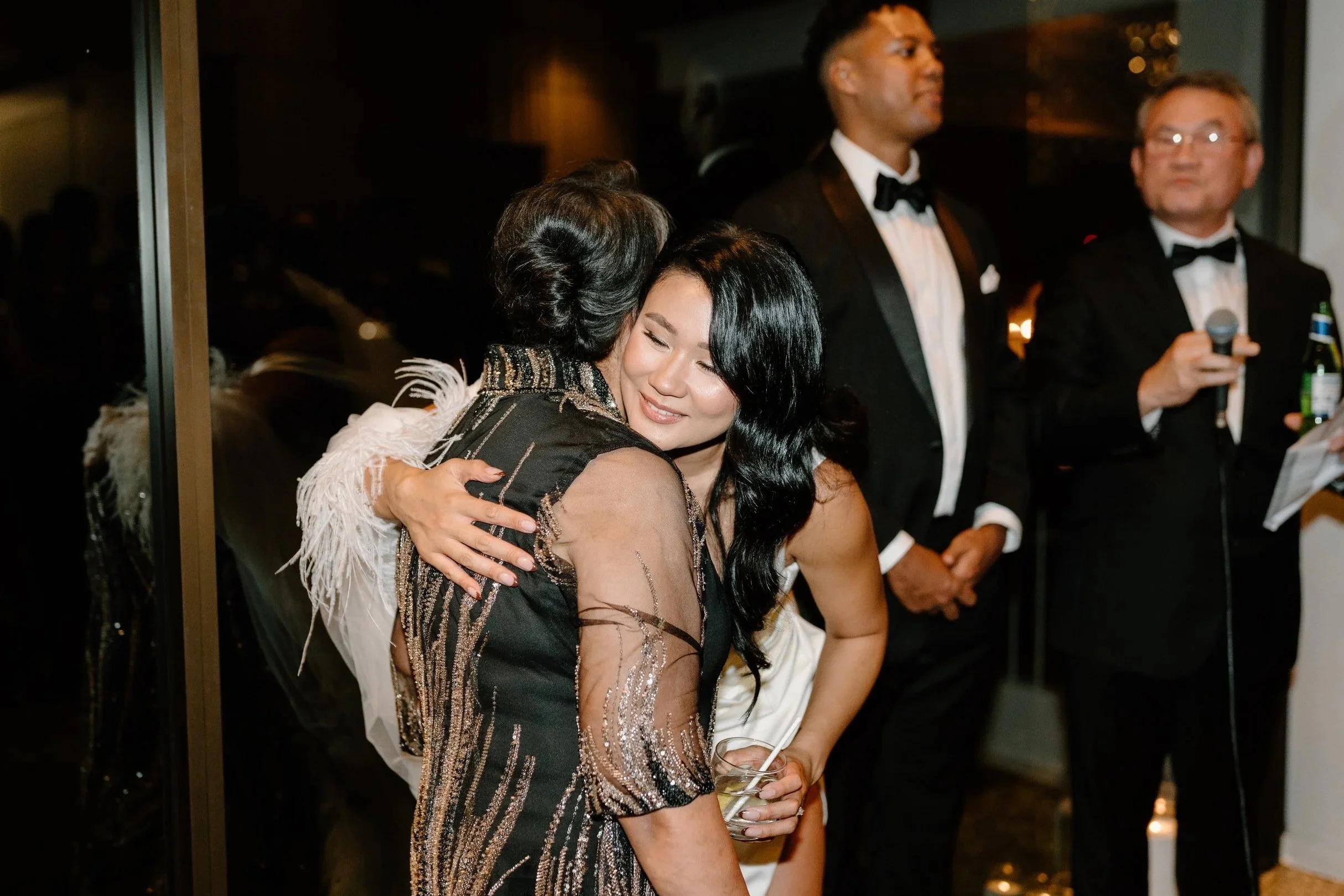 Two women hugging, one with dark, long hair in a black and gold dress, and the other in a white dress, at a formal event with men in tuxedos in the background.