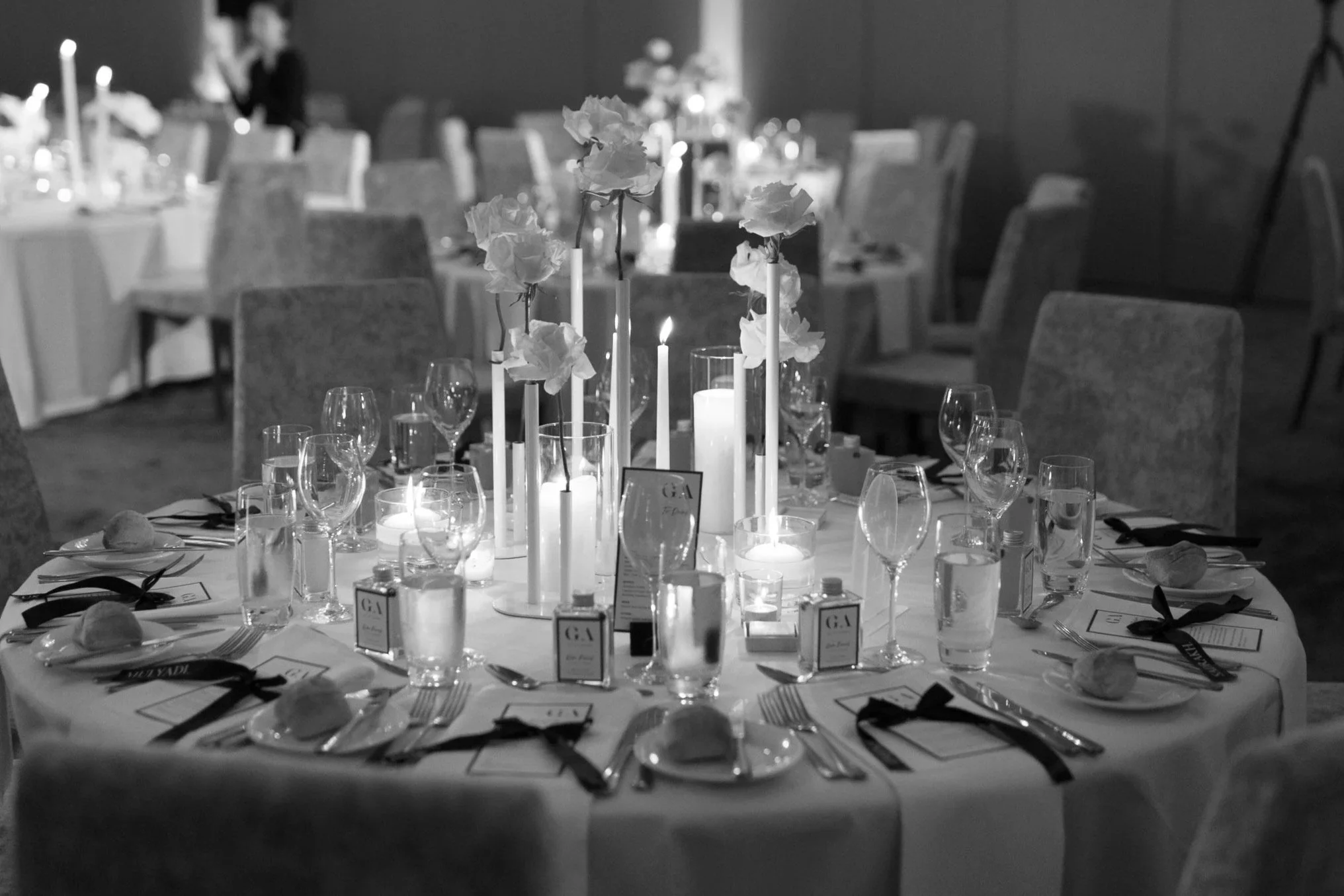 A round banquet table decorated with tall white candles, flowers in tall vases, wine glasses, water glasses, and black ribbons on white napkins, set for a formal event in a dimly lit room.