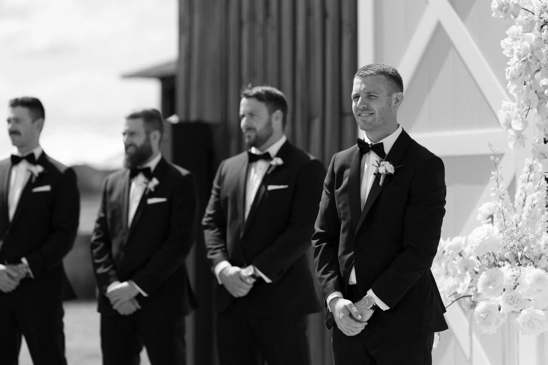 A man in a tuxedo standing during a wedding ceremony with four other men in tuxedos standing in a row outdoors.