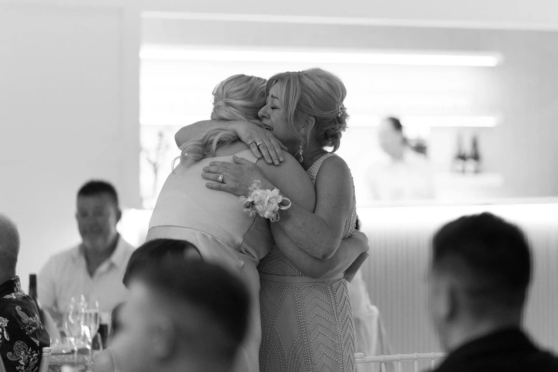 Two women embracing, one in a wedding dress, at a wedding reception, with guests seated at tables in the background.