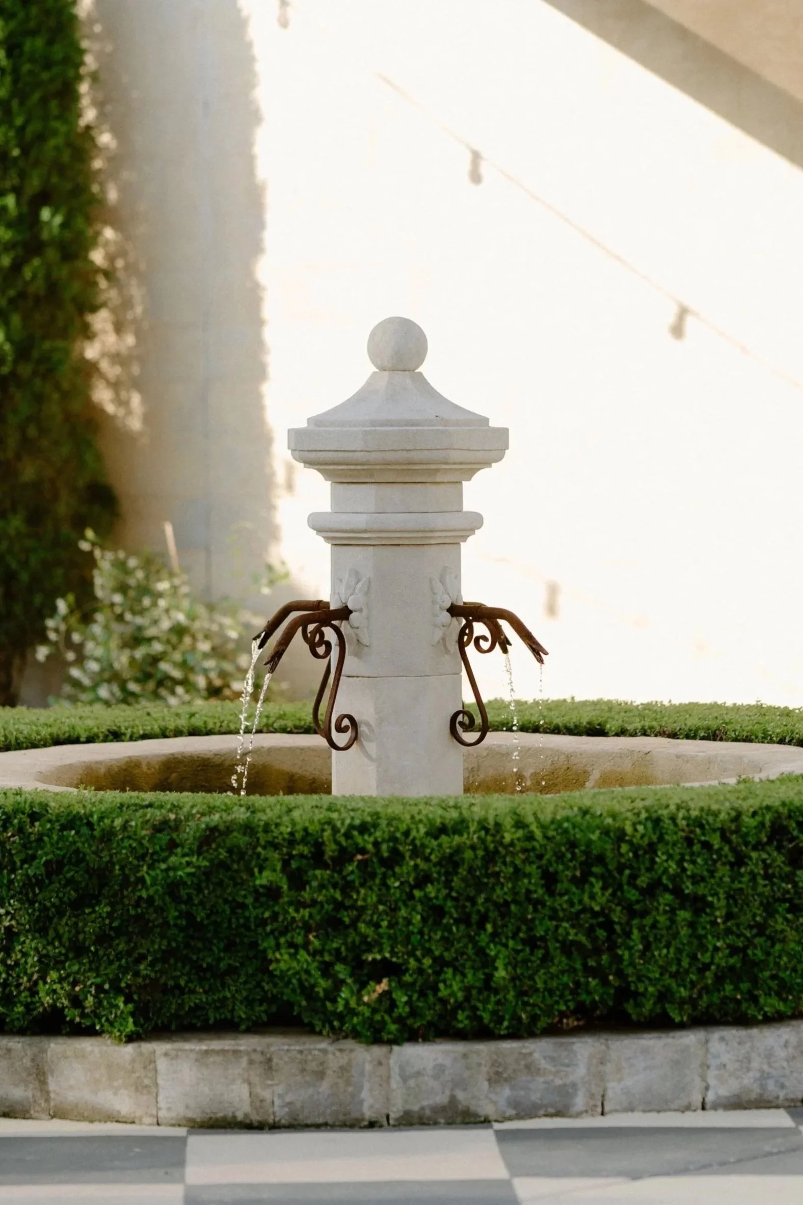 A small decorative fountain in an outdoor patio area, surrounded by a neatly trimmed hedge, with checkered black and white flooring, a covered patio roof, and a wall-mounted outdoor light.