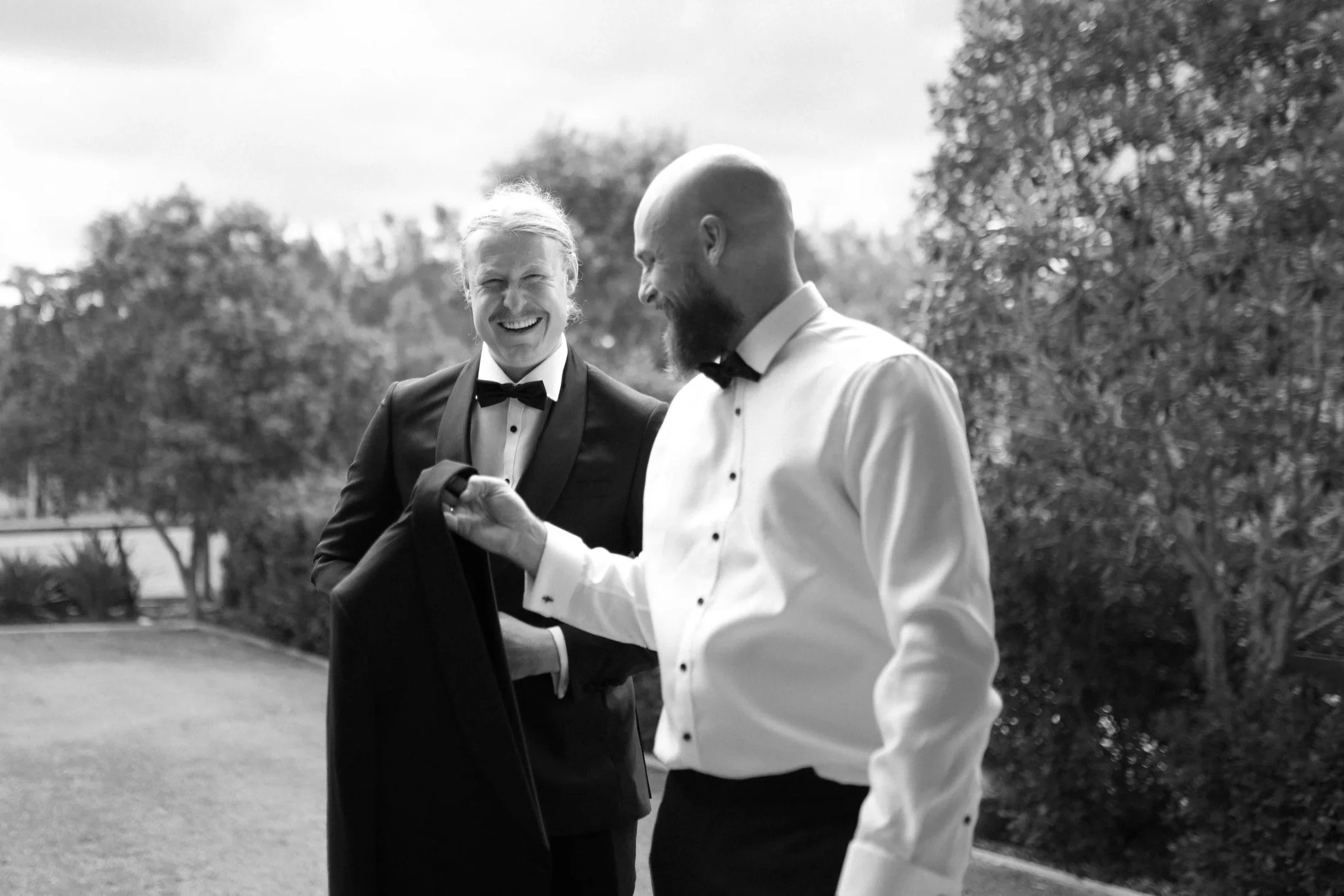 Two men in tuxedos, one with a bow tie and the other with a regular tie, are outdoors smiling and engaging in conversation. One man has a beard and a shaved head, the other has longer hair tied back. Trees and a cloudy sky are in the background.
