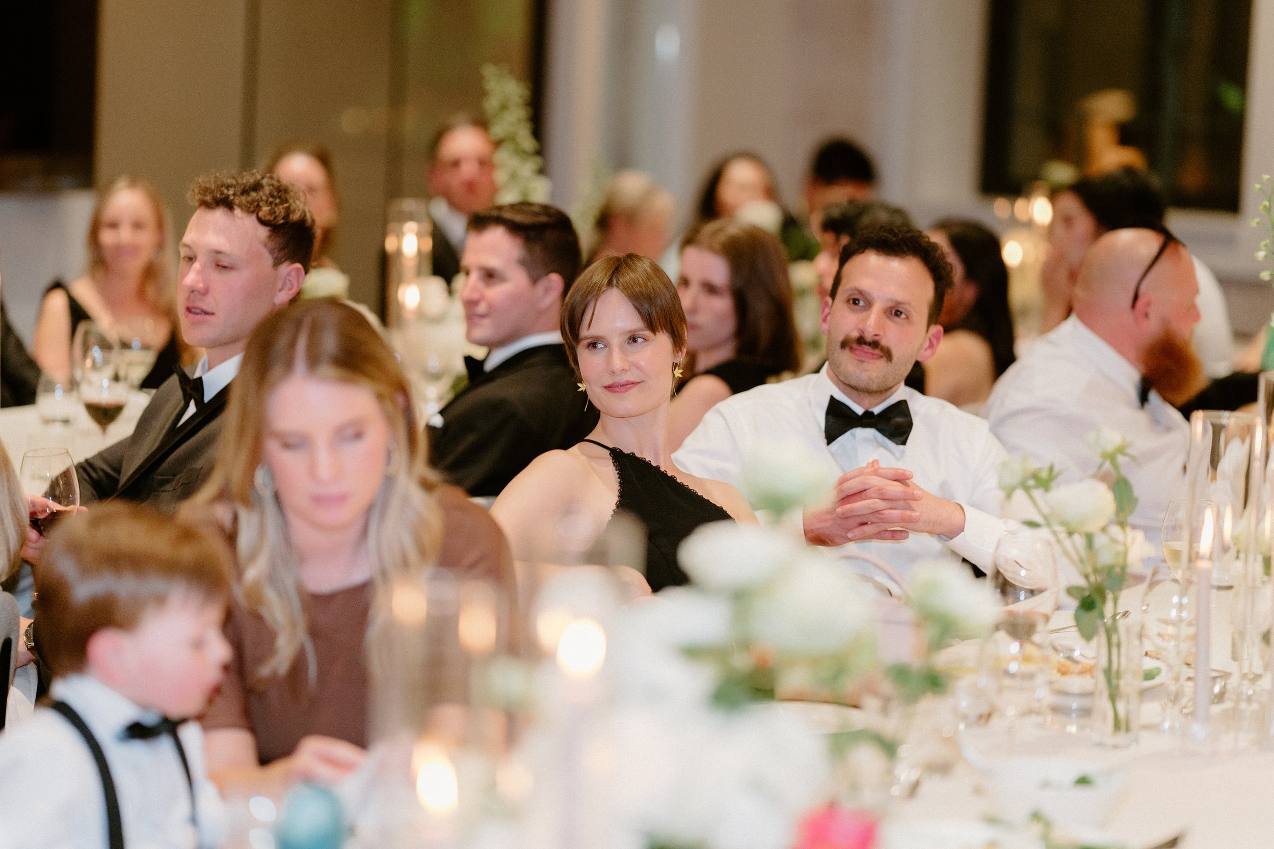 People sitting at a formal dinner table, dressed in tuxedos and evening gowns, attending a celebration or event, with candles and floral decorations on the table.